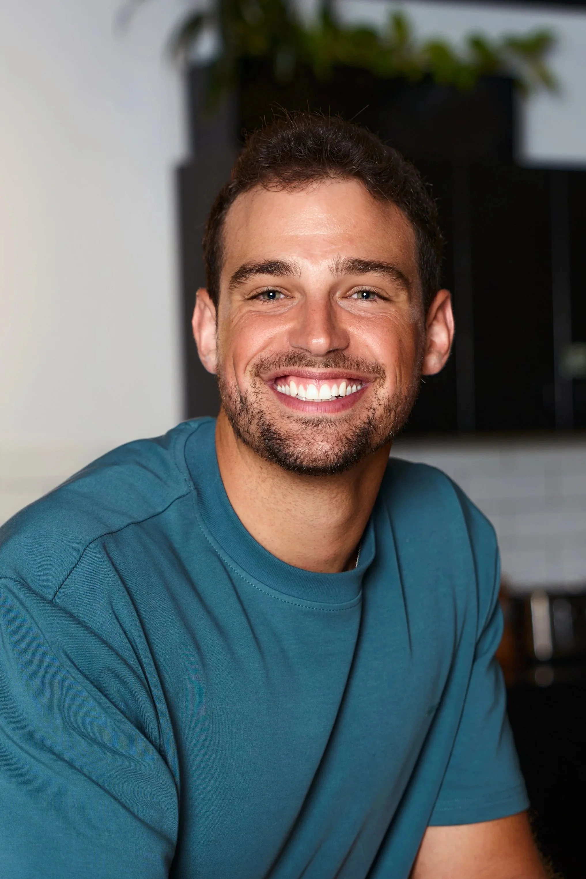 A woman with shoulder-length brown hair smiling, wearing a gray blazer over a white T-shirt, sitting indoors on a dark couch with a cozy ambiance.