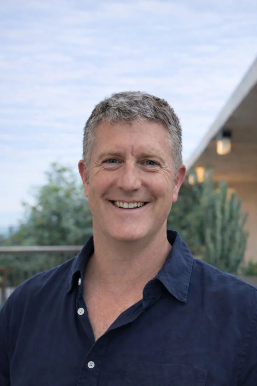 Smiling woman with shoulder-length blonde hair, wearing a black top and a necklace, indoors with warm lighting.