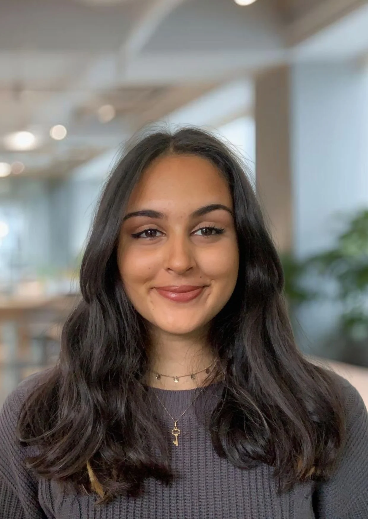 A woman with shoulder-length brown hair smiling, wearing a gray blazer over a white T-shirt, sitting indoors on a dark couch with a cozy ambiance.