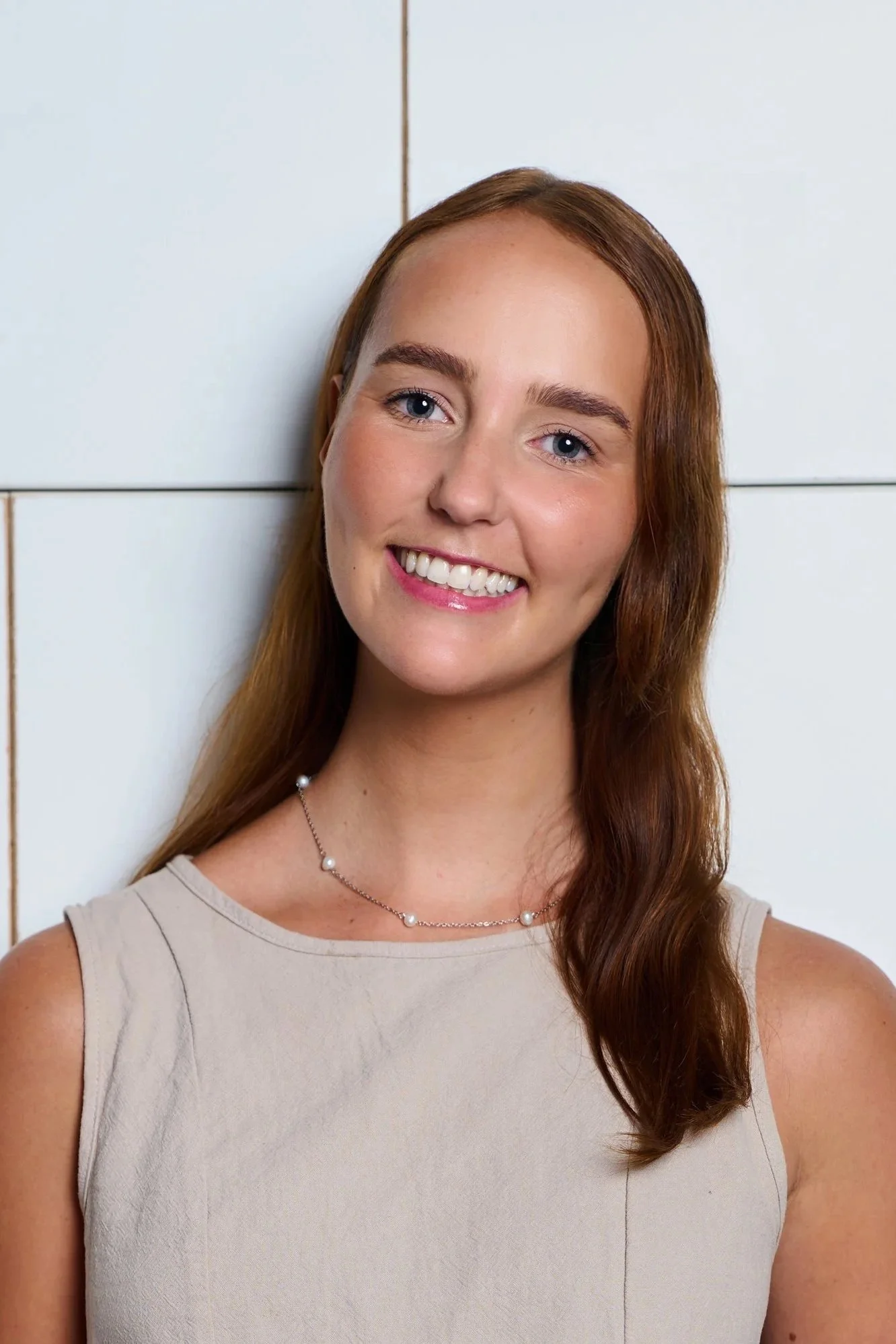 A woman with shoulder-length brown hair smiling, wearing a gray blazer over a white T-shirt, sitting indoors on a dark couch with a cozy ambiance.