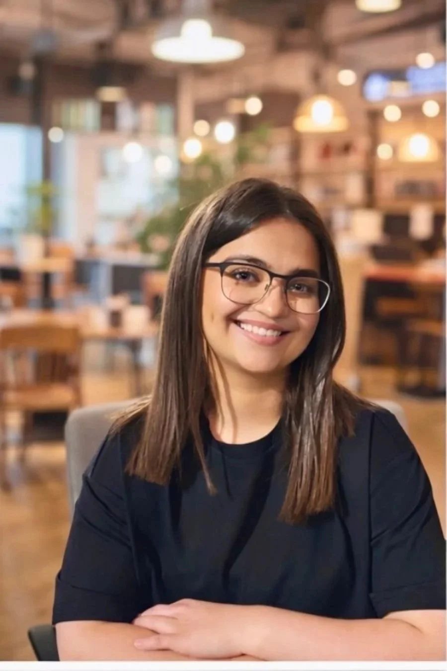Smiling woman with shoulder-length brown hair, wearing a black top and a glasses, indoors in an office space.
