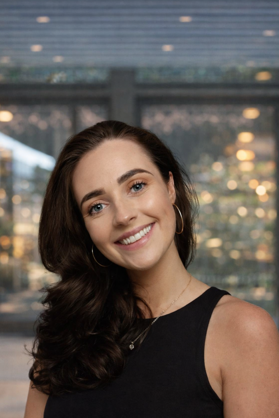 A woman with long dark hair wearing a navy sleeveless top, gold hoop earrings, a gold necklace, and glasses resting on her head, smiling while sitting on a wooden bench with pink cushions in the background.