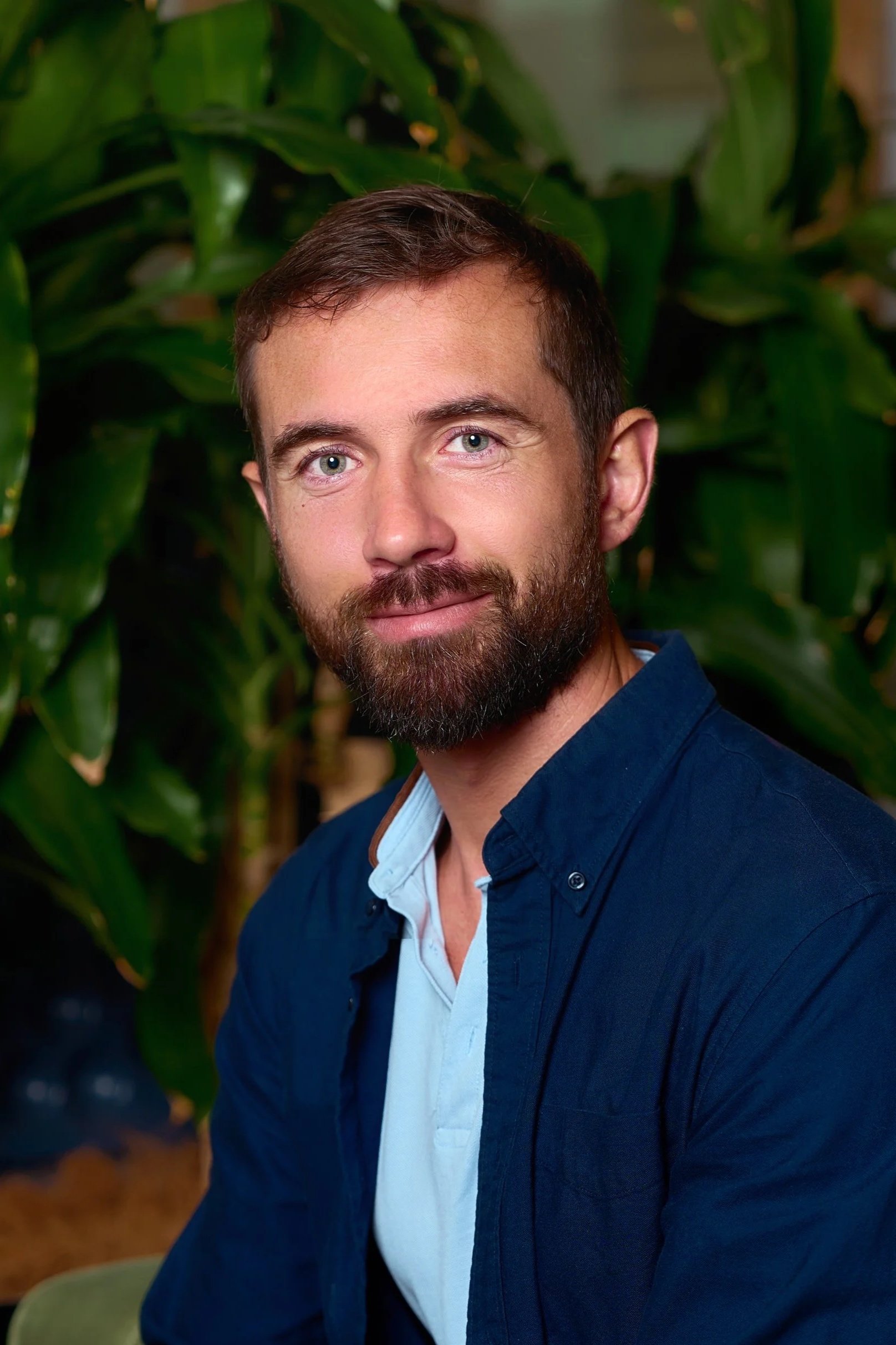 A young woman smiling and sitting at a wooden desk with her hand resting on her chin, in front of a silver HP laptop.