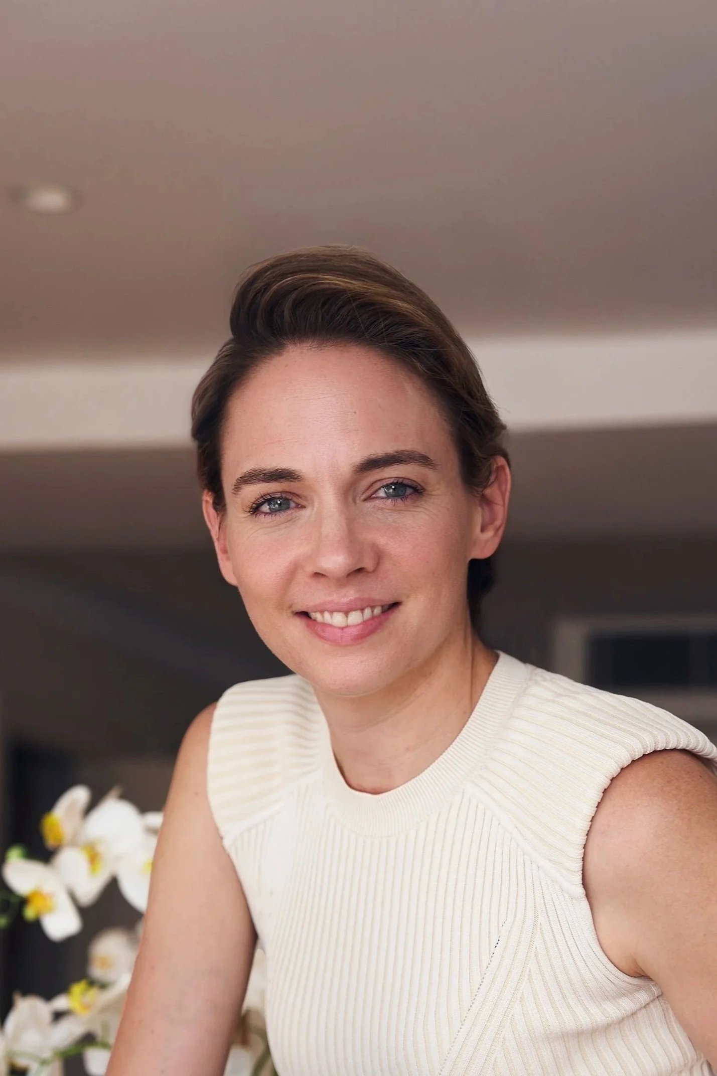 A woman with short brown hair smiling and standing with arms crossed, wearing a black blazer with gold buttons in front of beige curtains.