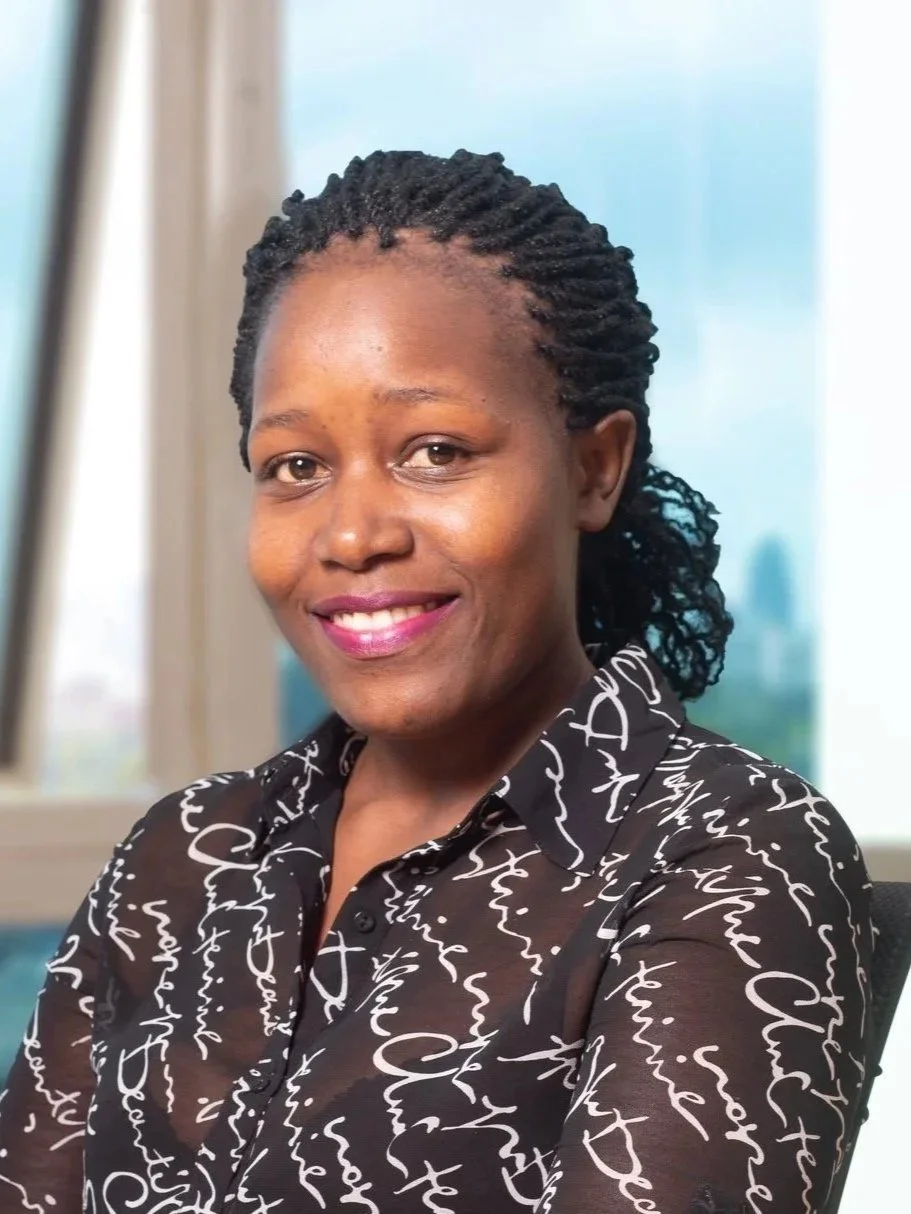 Smiling woman with shoulder-length blonde hair, wearing a black top and a necklace, indoors with warm lighting.