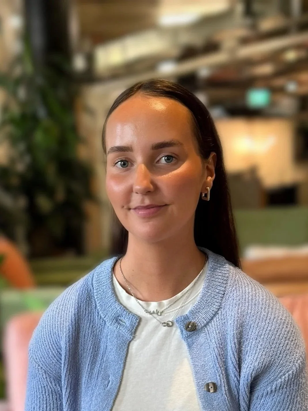 A woman with shoulder-length brown hair smiling, wearing a gray blazer over a white T-shirt, sitting indoors on a dark couch with a cozy ambiance.
