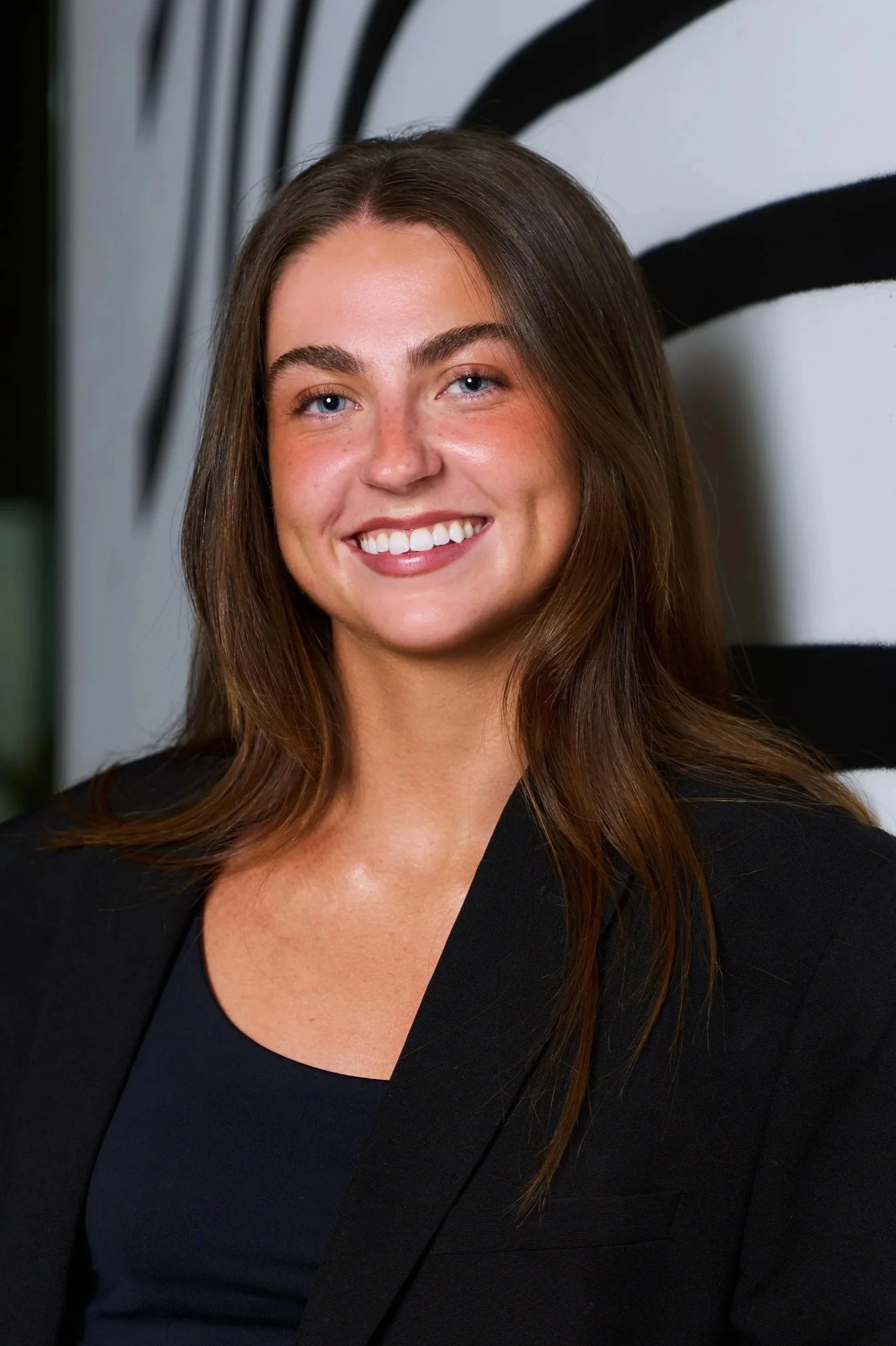 A woman with shoulder-length brown hair smiling, wearing a gray blazer over a white T-shirt, sitting indoors on a dark couch with a cozy ambiance.
