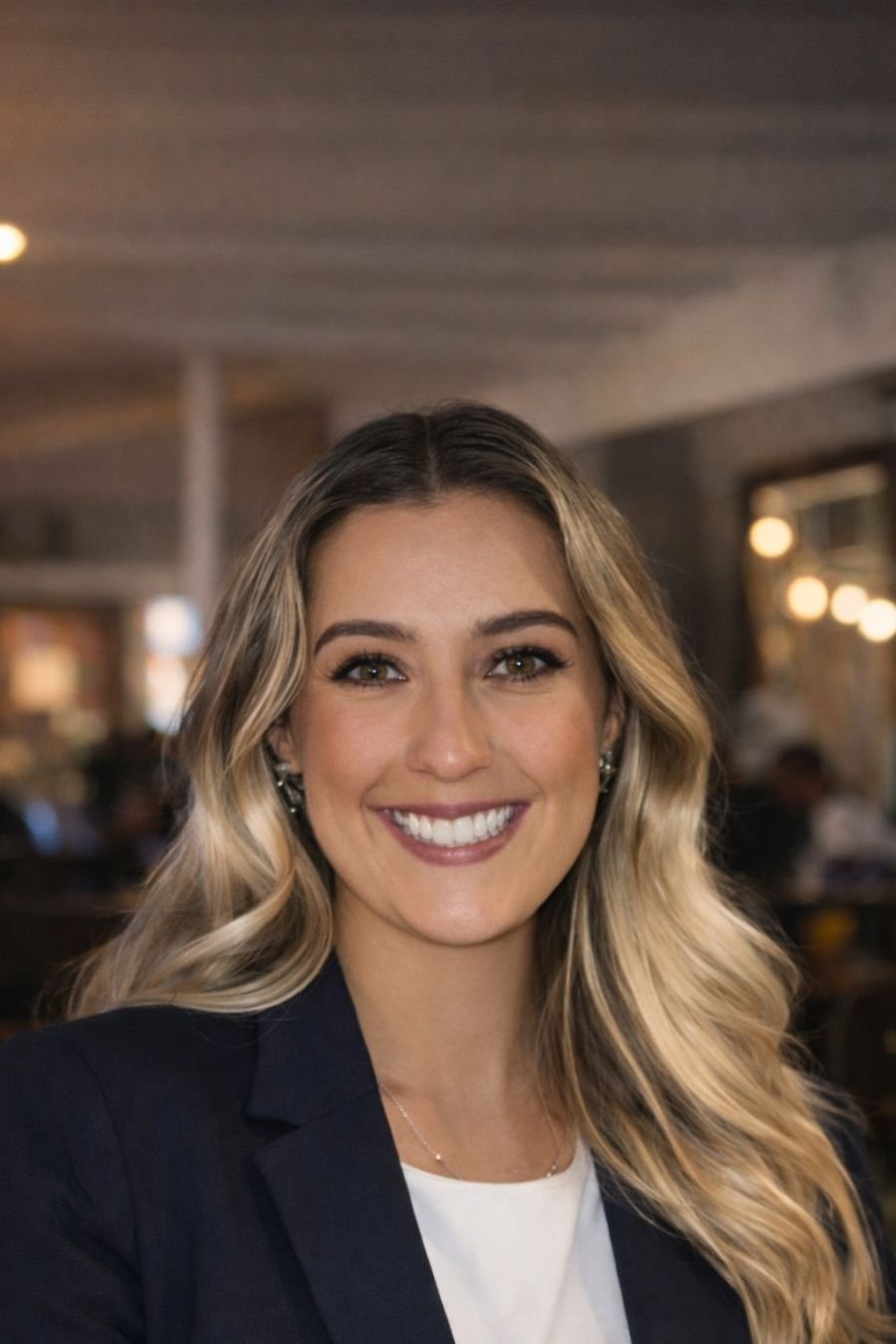 A woman with shoulder-length brown hair smiling, wearing a gray blazer over a white T-shirt, sitting indoors on a dark couch with a cozy ambiance.