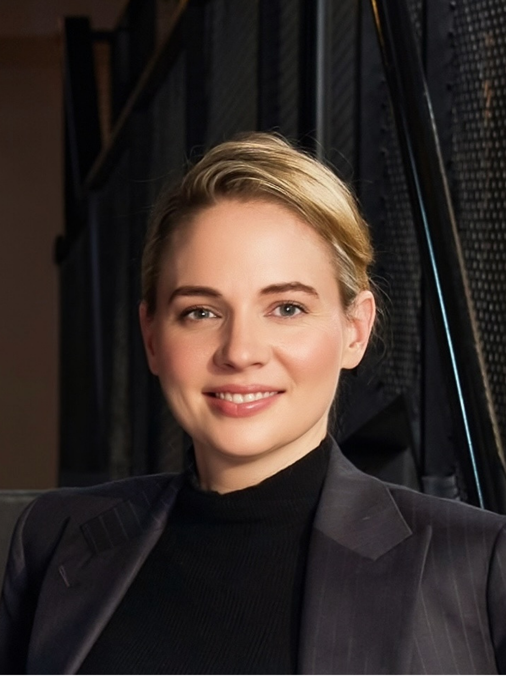A woman with short brown hair smiling and standing with arms crossed, wearing a black blazer with gold buttons in front of beige curtains.