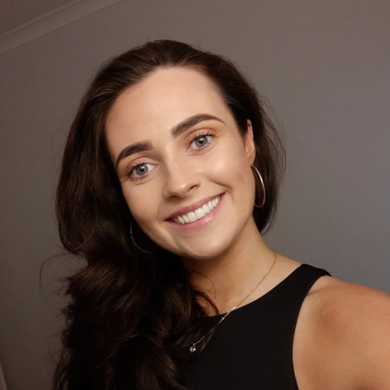 A woman with long dark hair wearing a navy sleeveless top, gold hoop earrings, a gold necklace, and glasses resting on her head, smiling while sitting on a wooden bench with pink cushions in the background.