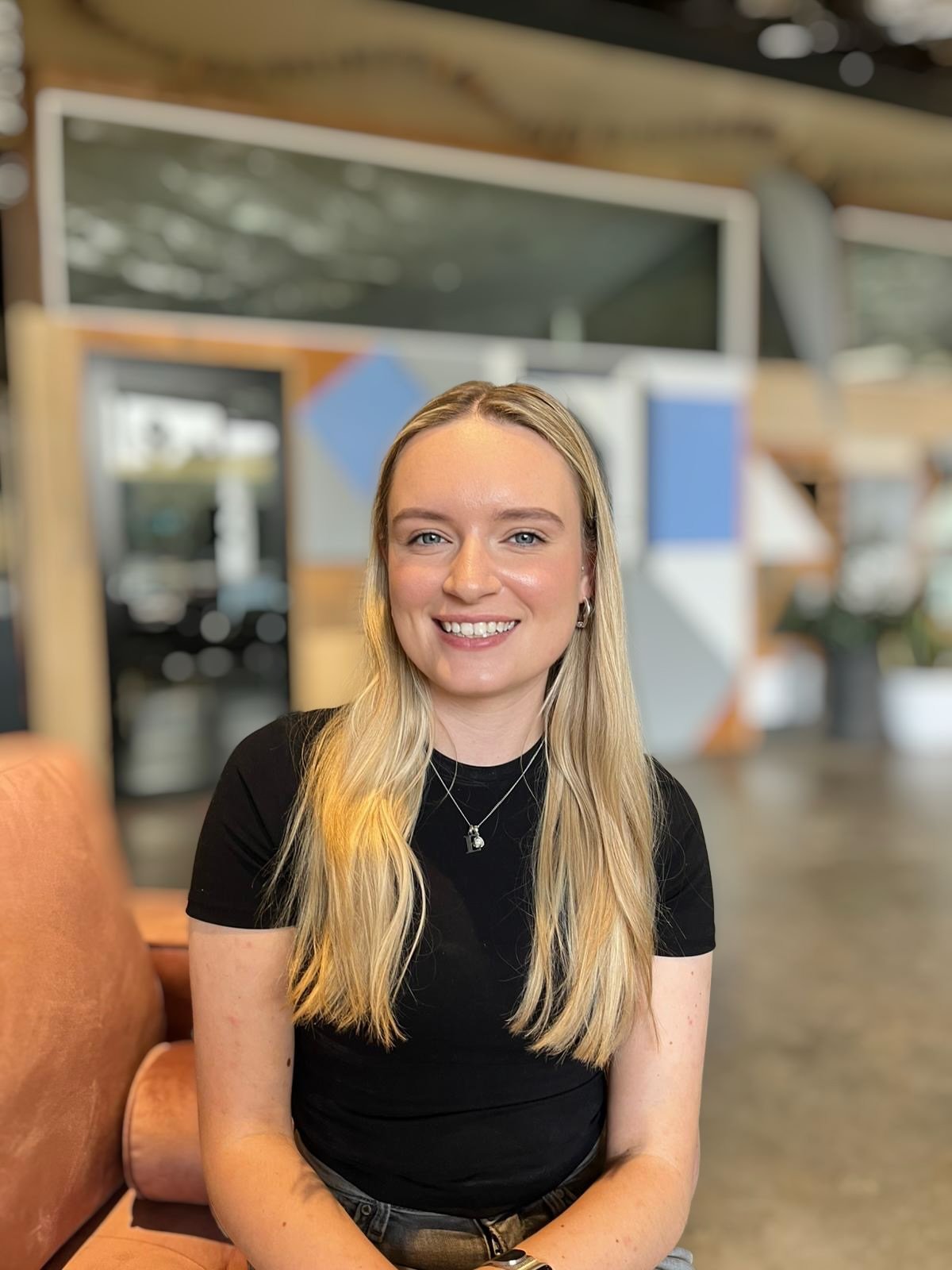 Smiling woman with shoulder-length blonde hair, wearing a black top and a necklace, indoors with warm lighting.