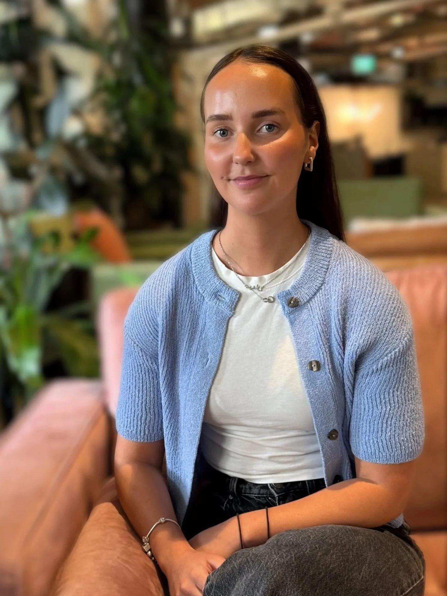 Young woman with shoulder-length dark hair, smiling, wearing a black top, sitting with her chin resting on her hand in a well-lit room.