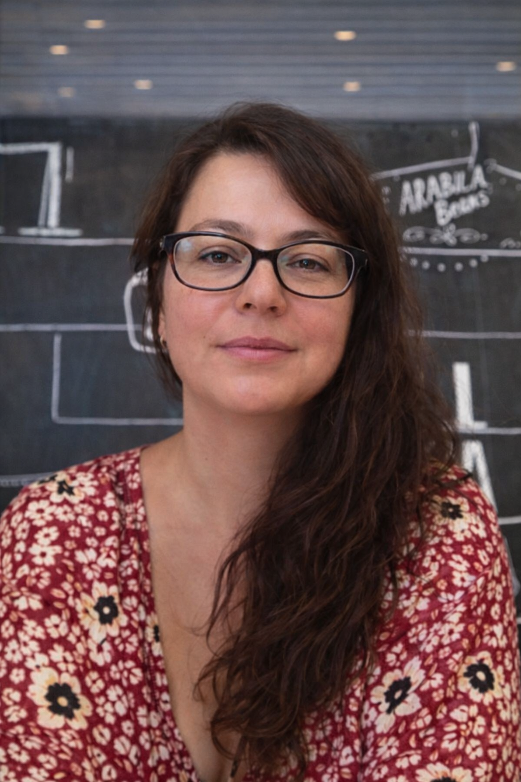 Smiling woman with shoulder-length blonde hair, wearing a black top and a necklace, indoors with warm lighting.