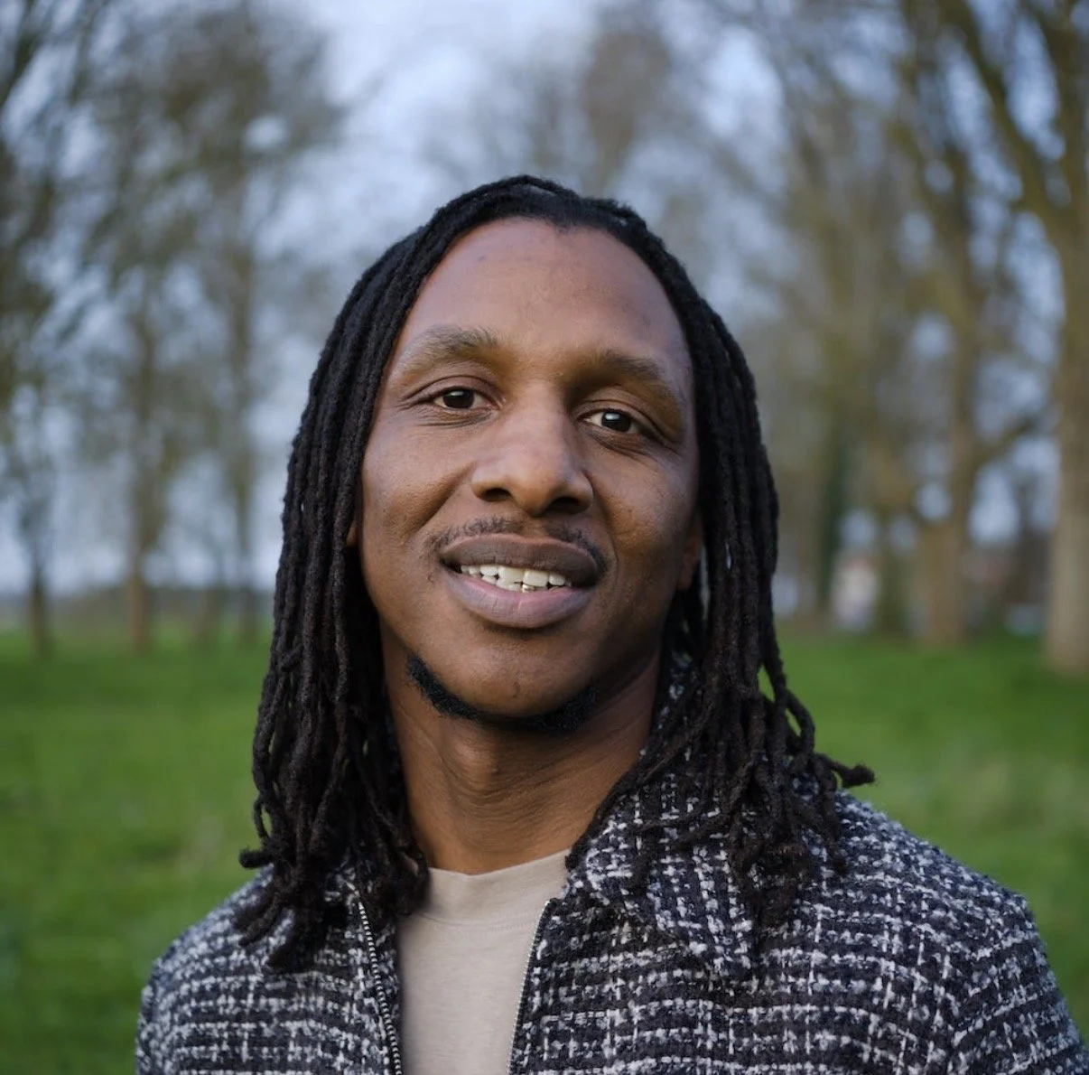 A man with long dreadlocks smiling outdoors with trees in the background.