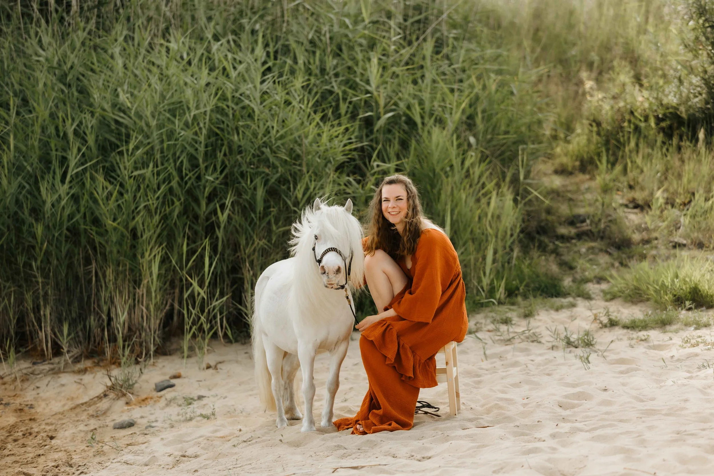 Frau in einem rostfarbenen Boho Kleid mit Schlitz sitzt auf einem Hocker. Daneben ein weißes Shetlandpony an der Elbe