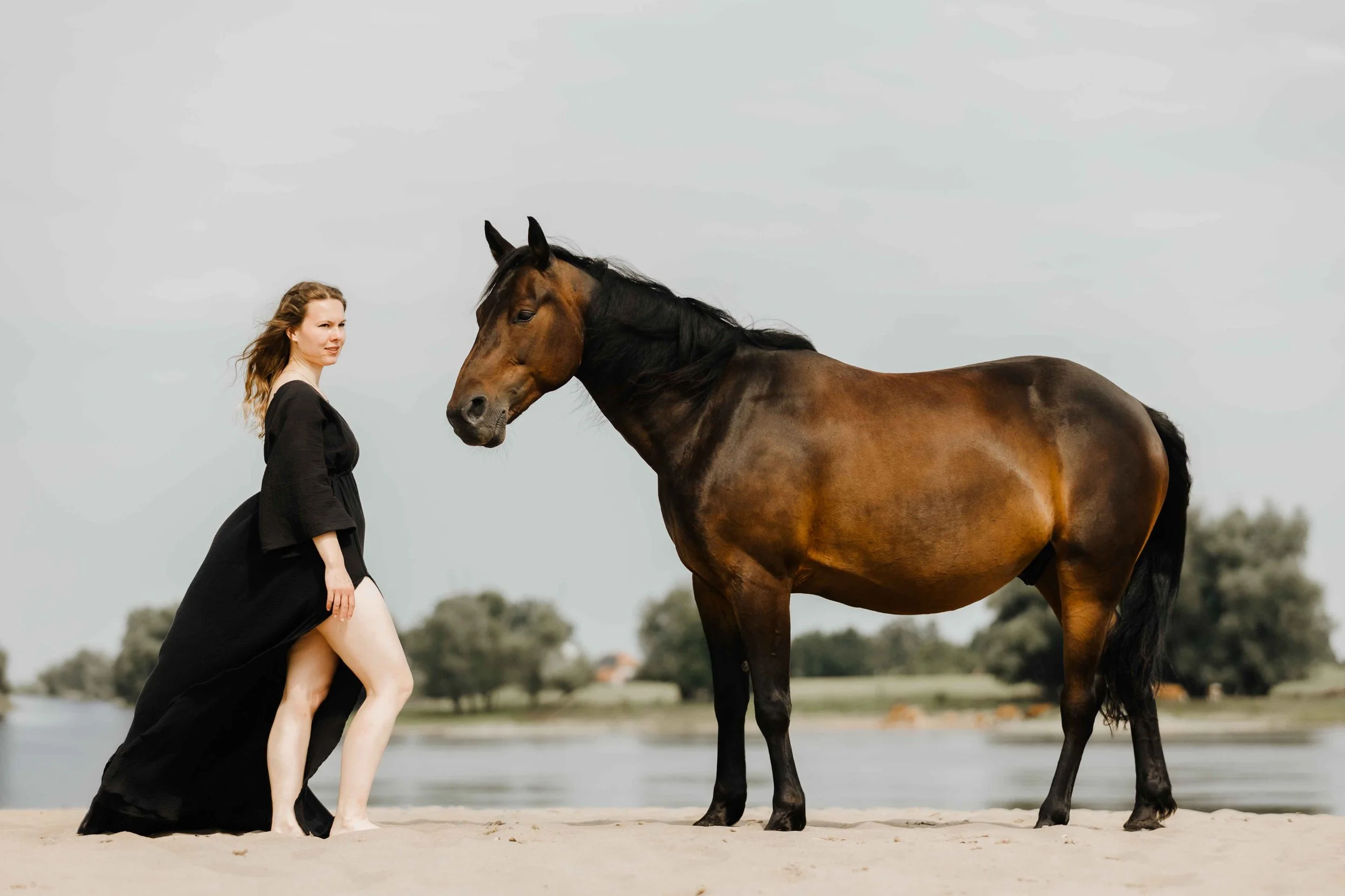 Frau in einem eleganten schwarzen Kleid mit ihrem Pferd an der Elbe