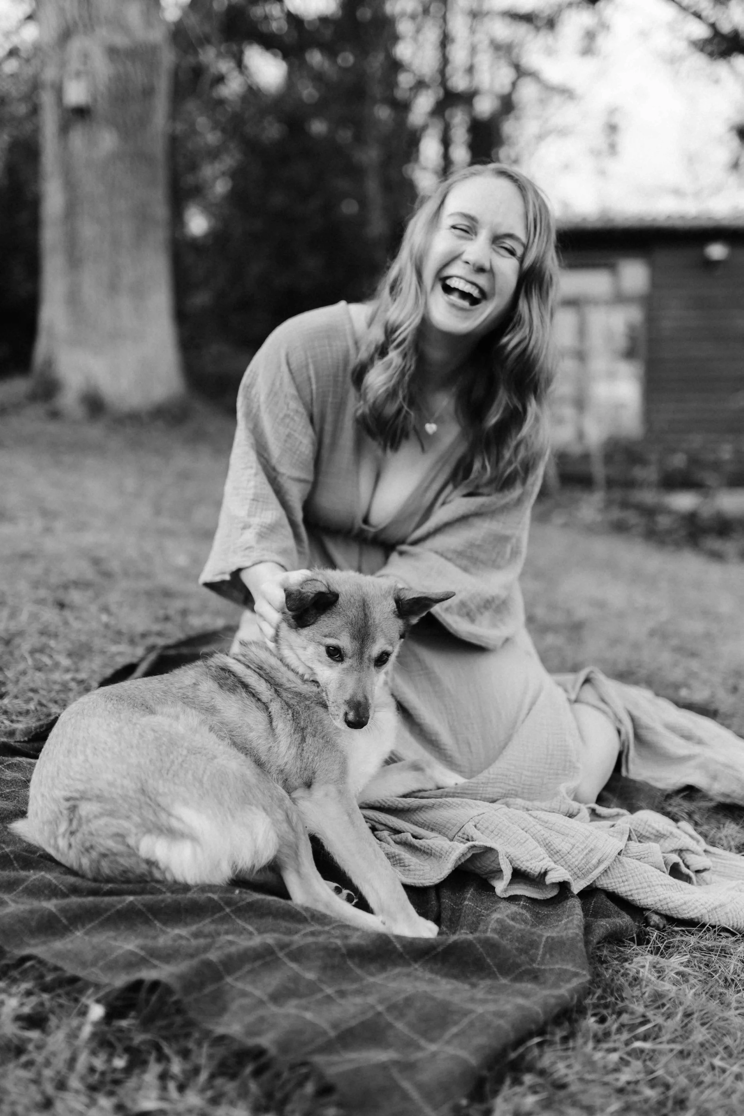 Schwarz-weiß Aufnahme von einer Frau in einem Kleid die aus ganzem Herzen lacht. Vor ihr liegt ihr Hund auf einer Decke und sie streichelt ihn liebevoll.