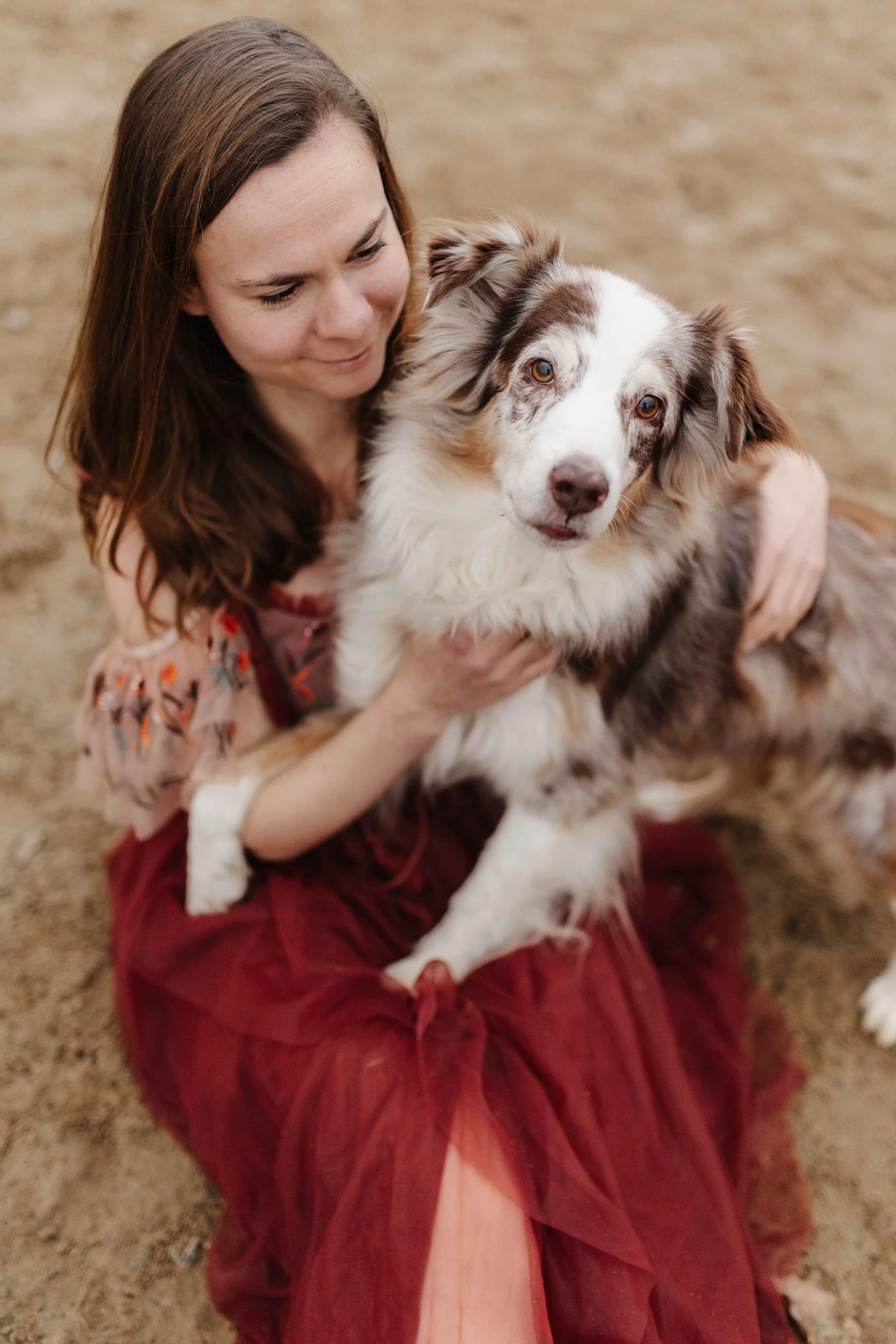 Frau in eine schönen roten Blumenkleid sitzt auf dem Boden eines Reitplatzes. Auf ihrem Schoß liegt ein Australian Shepherd in Red Merle.