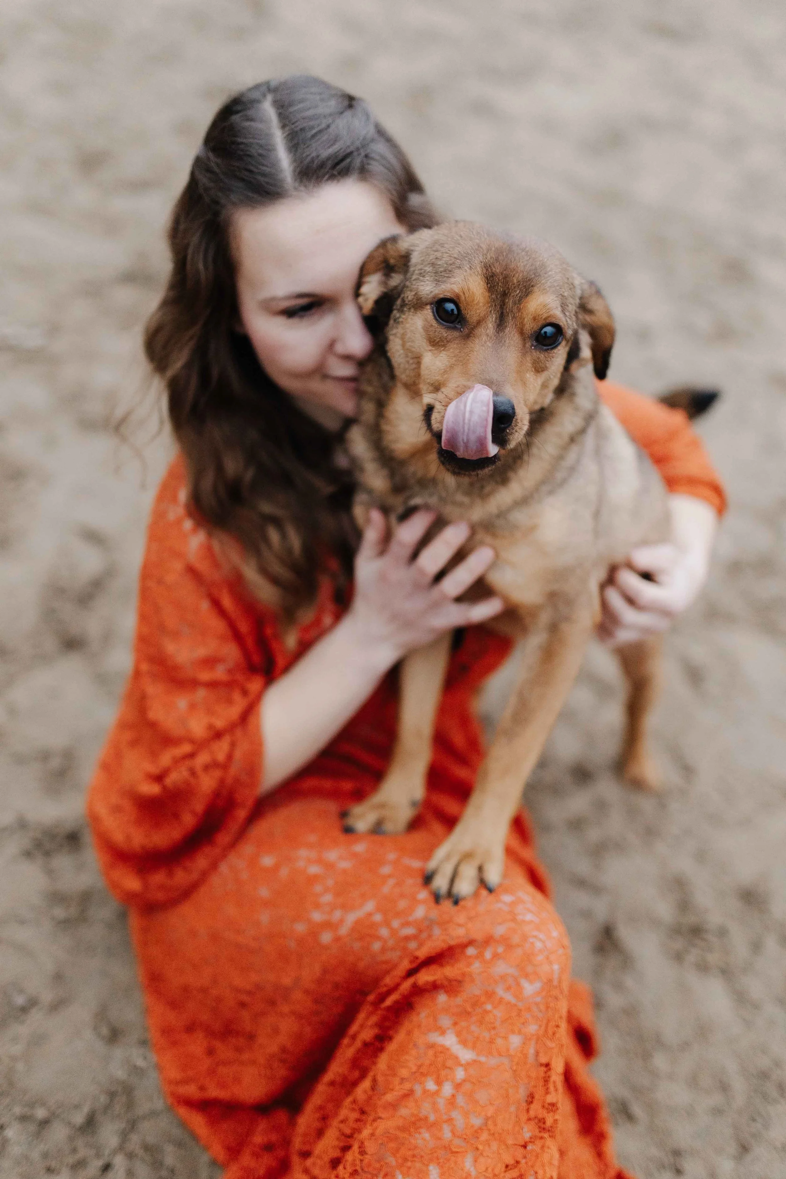 Frau im rostfarbenen Spitzekleid sitzt auf dem Boden eines Reitplatzes und auf ihrem Schoß steht ein brauner Hund, der sich über seine Schnauze leckt.