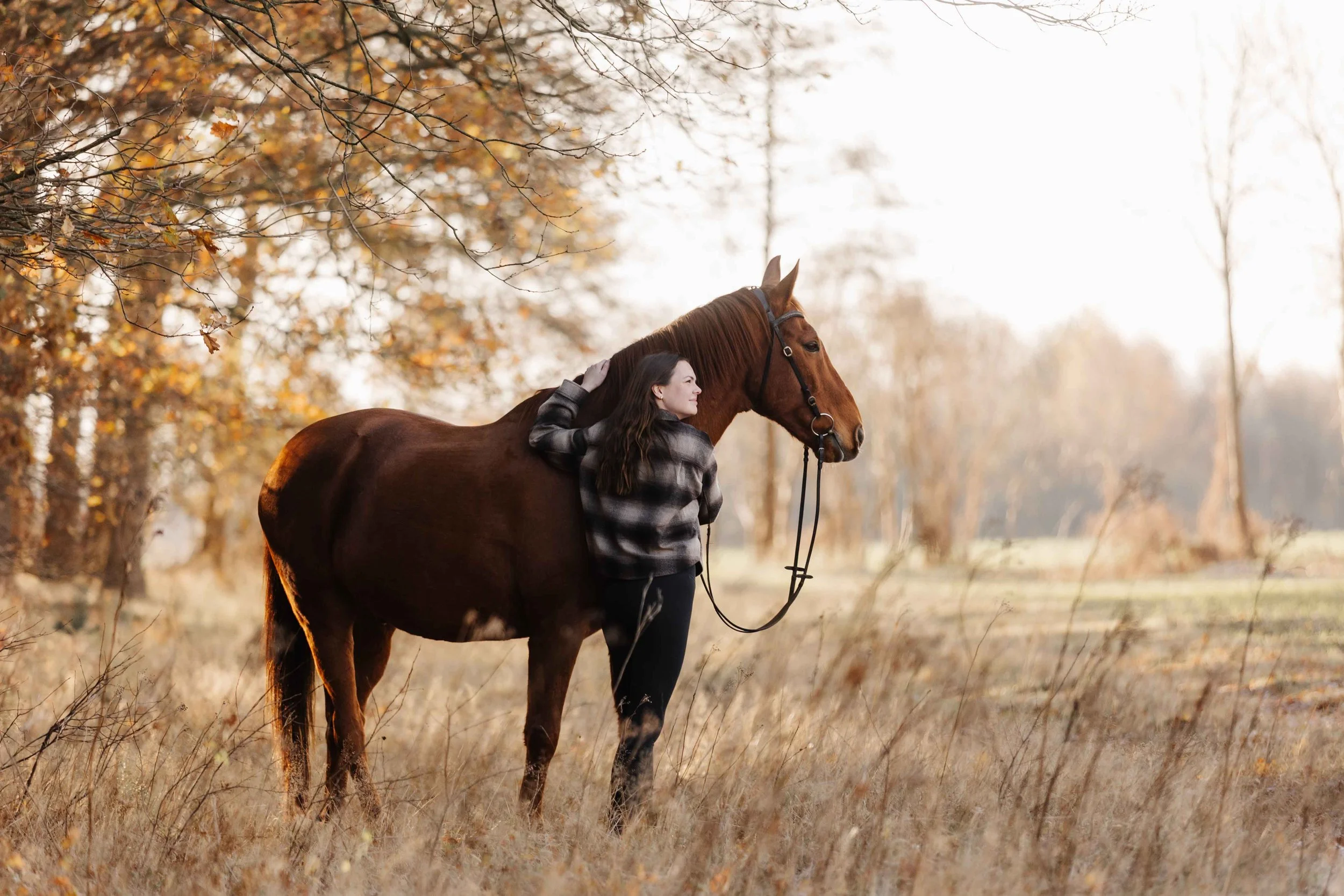 Eine Frau steht eng an einem fuchsfarbenen Pferd in einer herbstlichen Wiese, streichelt seinen Hals und blickt lächelnd in die warme Nachmittagssonne