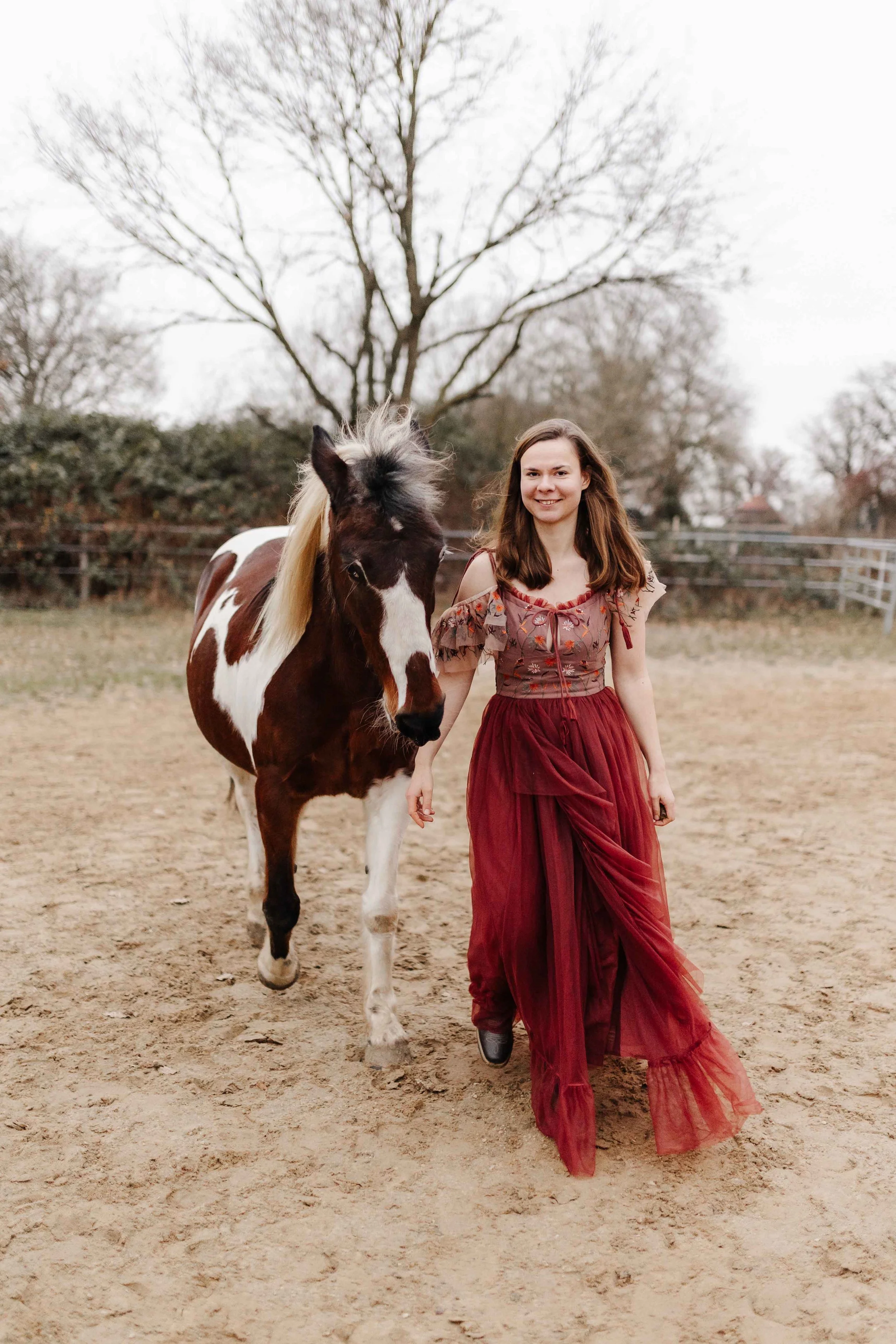 Frau im roten Blumenkleid geht auf einem Reitplatz entlang. Daneben ein geschecktes Pferd