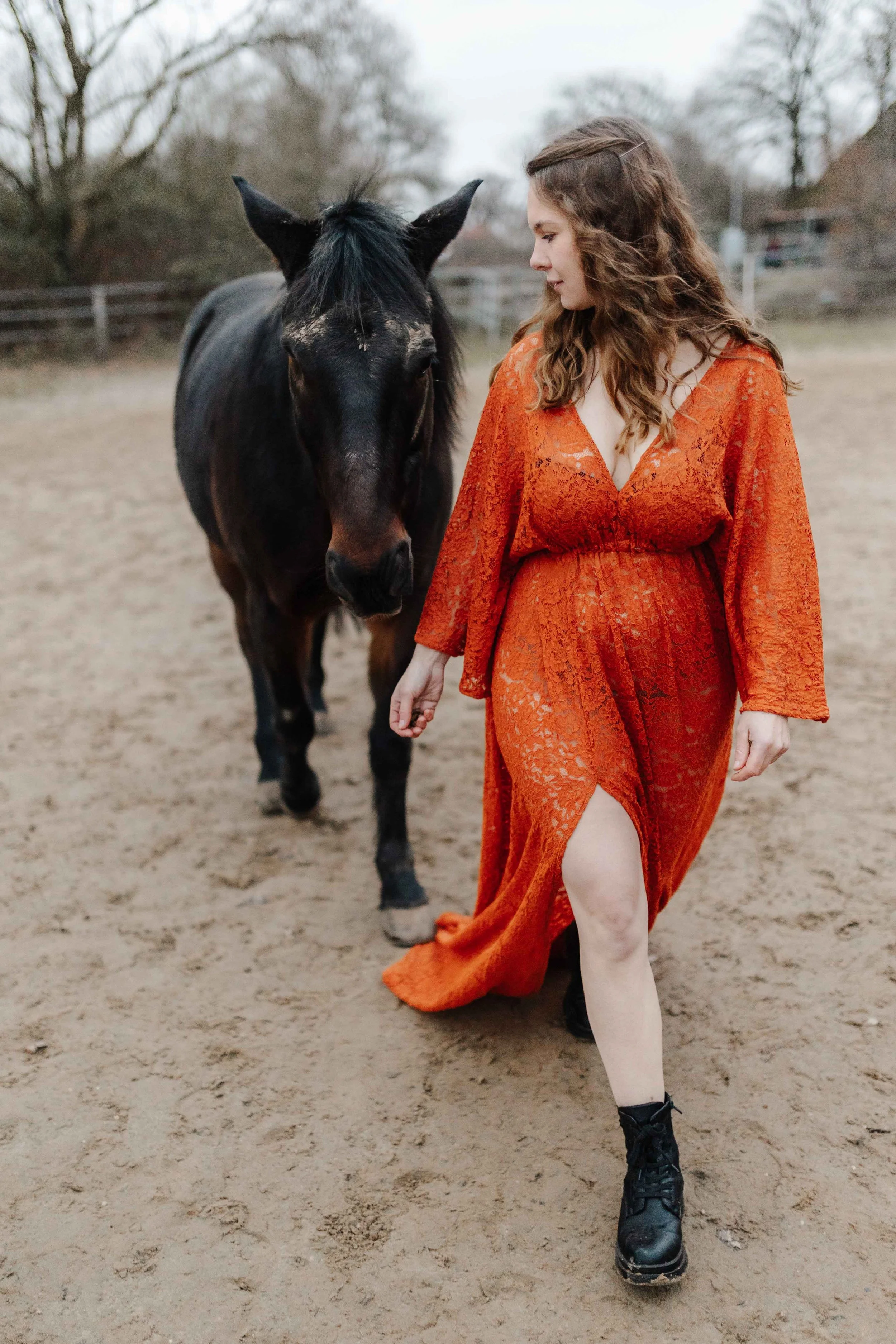 Eine Frau in einem rostfarbenen Spitzekleid mit Schlitz geht über den Reitplatz. Hinter iher ist ein schwarzes Pferd das ihr folgt.