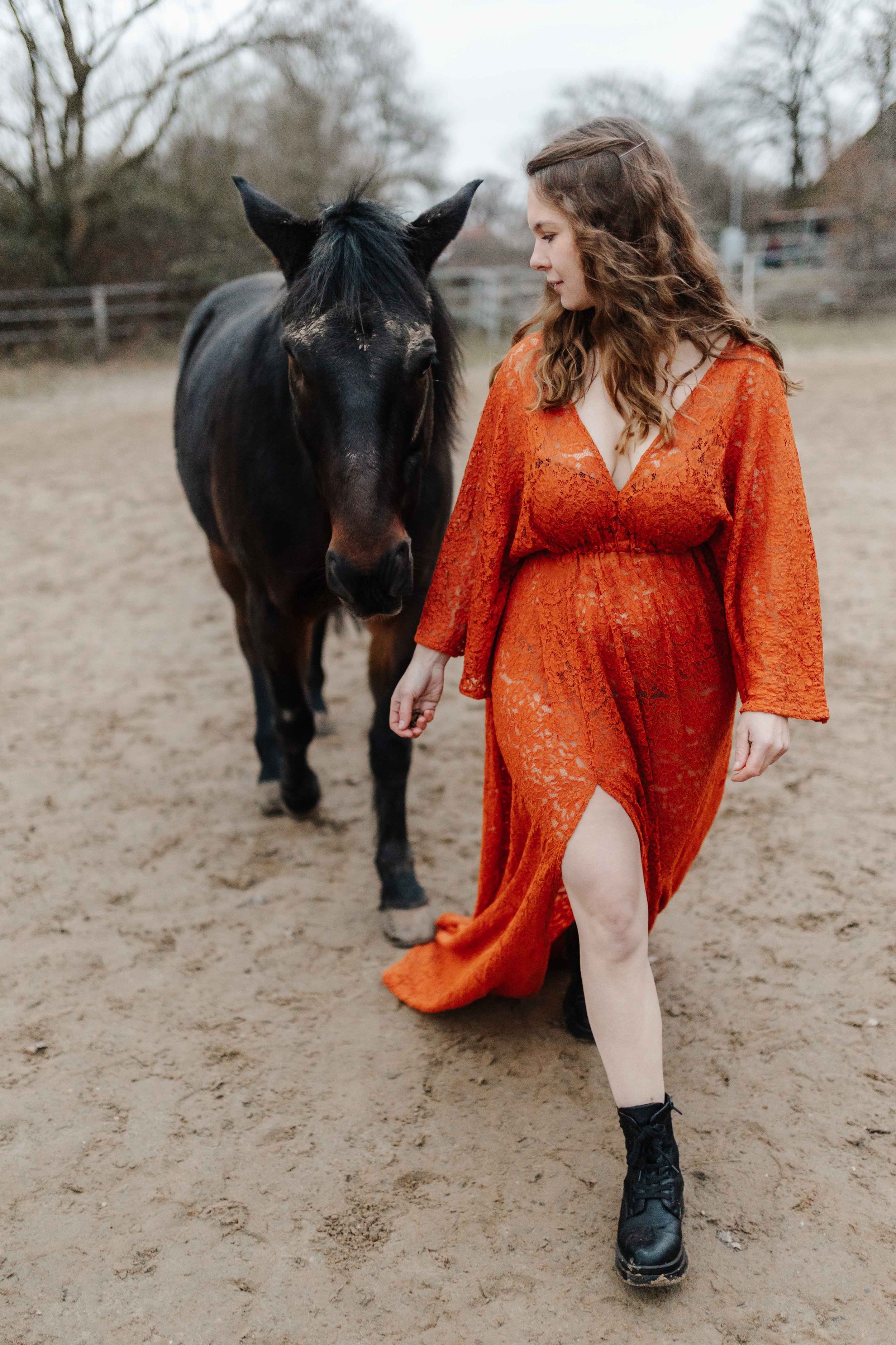 Frau mit rostfarbenen Spitzenkleid mit Schlitz geht mit ihrem Pferd auf einem Reitplatz