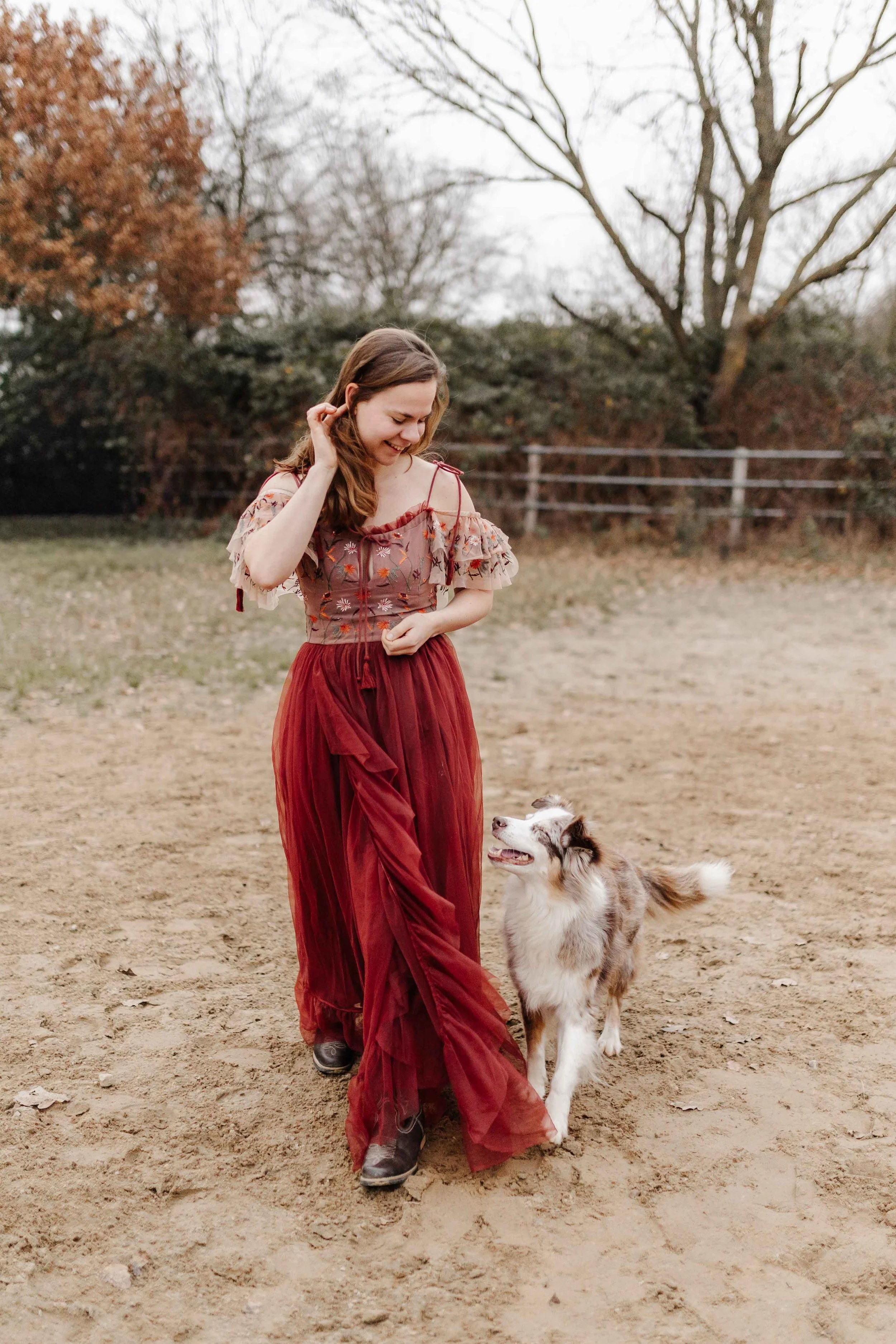 Frau im roten Blumenkleid geht auf einem Reitplatz entlang. Daneben läuft ein Australian Shepherd und schaut sie aufmerksam an