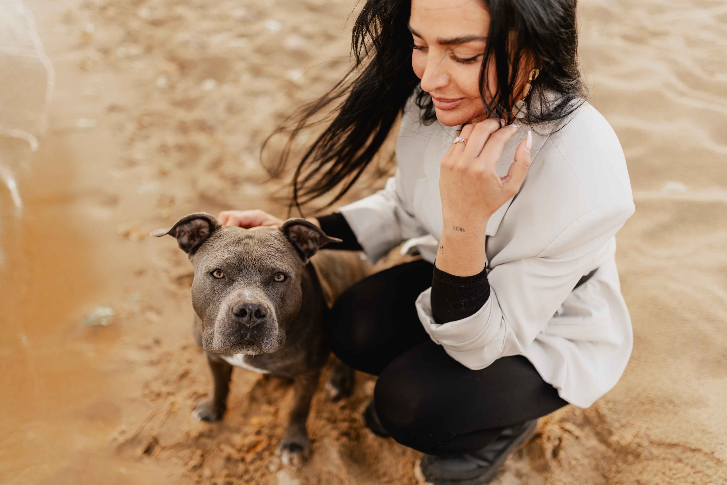Frau hockt am sandigen Seeufer und streichelt ihren grauen Hund, der aufmerksam in die Kamera schaut