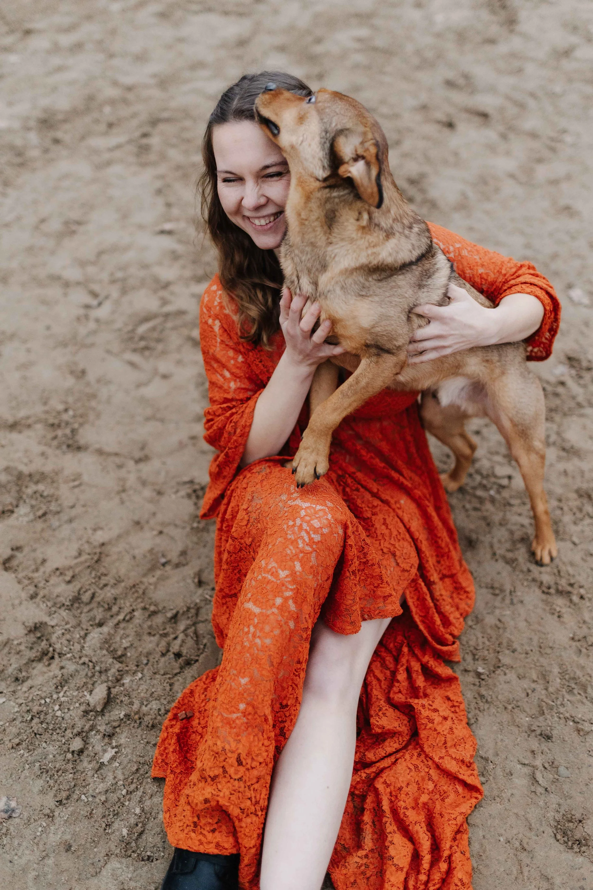 Auf dem Boden eines Reitplatzes sitzt eine Frau in einem rostfarbenen Spitzekleid. Auf ihrem Schoß steht ein brauner Hund der ihr übers Gesicht schleckt.