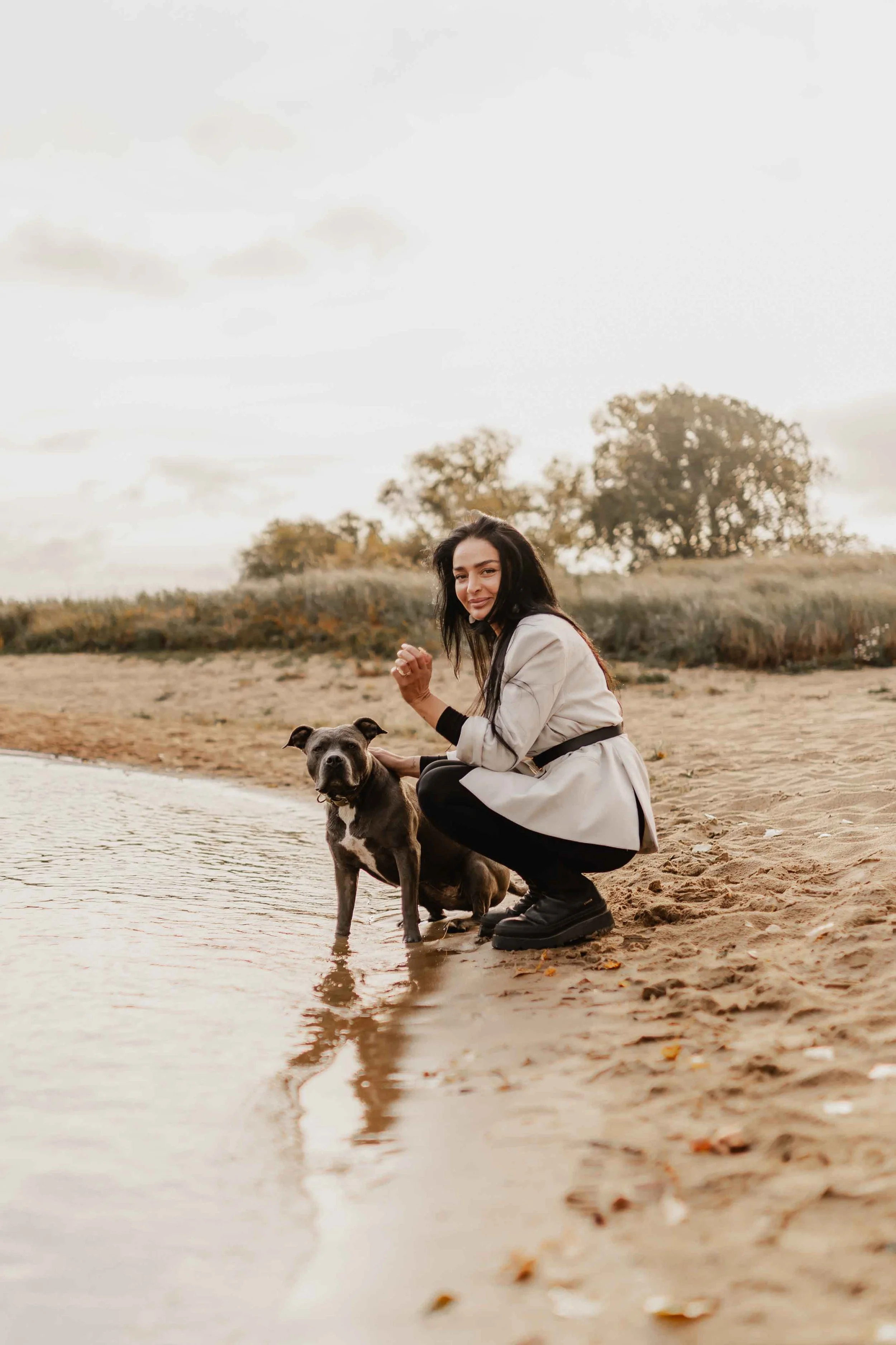 Frau hockt am Ufer eines Sees neben ihrem grauen Hund, beide blicken ruhig in die Kamera, sanftes Licht und herbstliche Stimmung