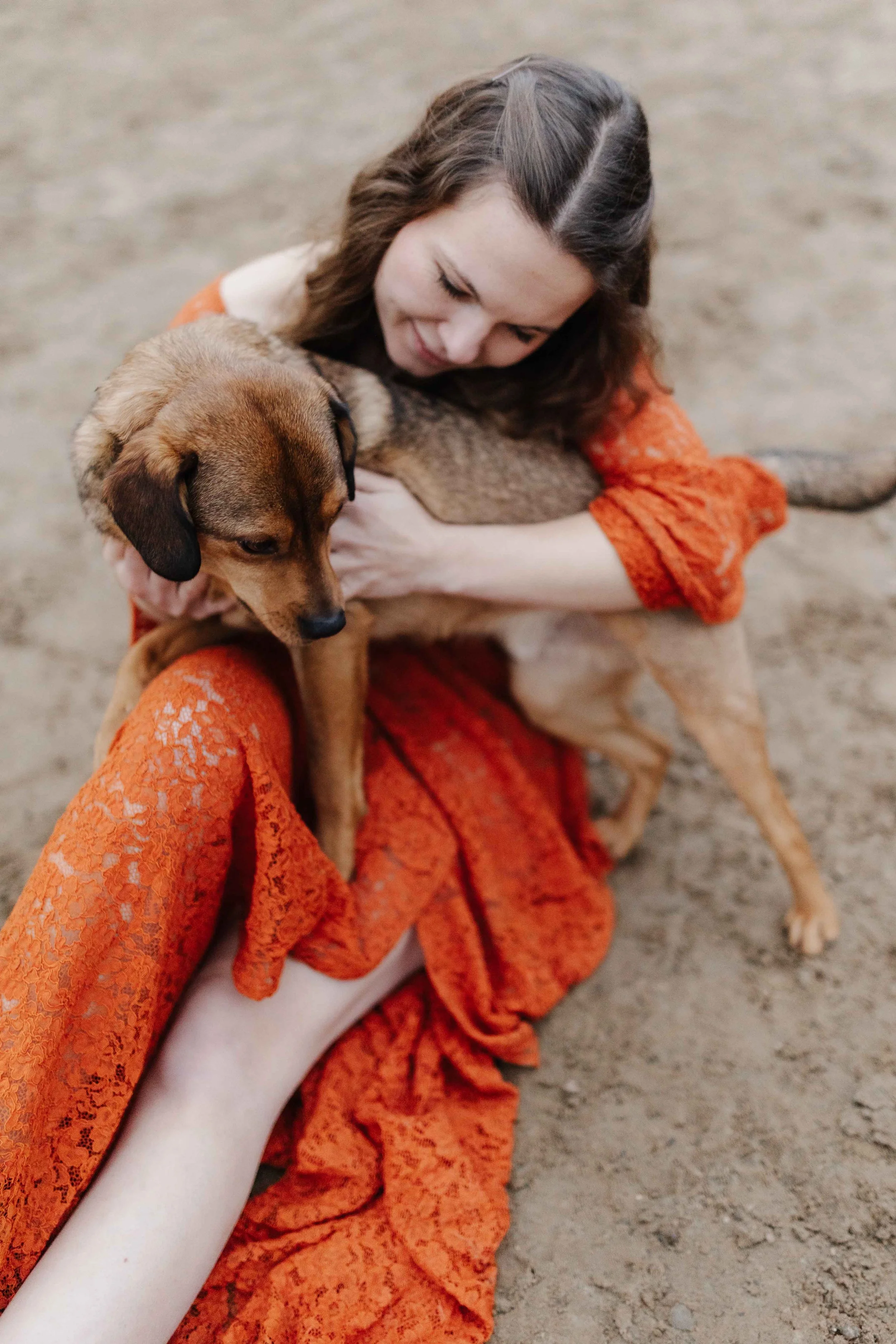 Frau in einem rostfarbenen Spitzekleid sitzt auf dem Boden eines Reitplatzes. Auf ihrem Schoß steht ein brauner Hund der sich an die Frau kuschelt.