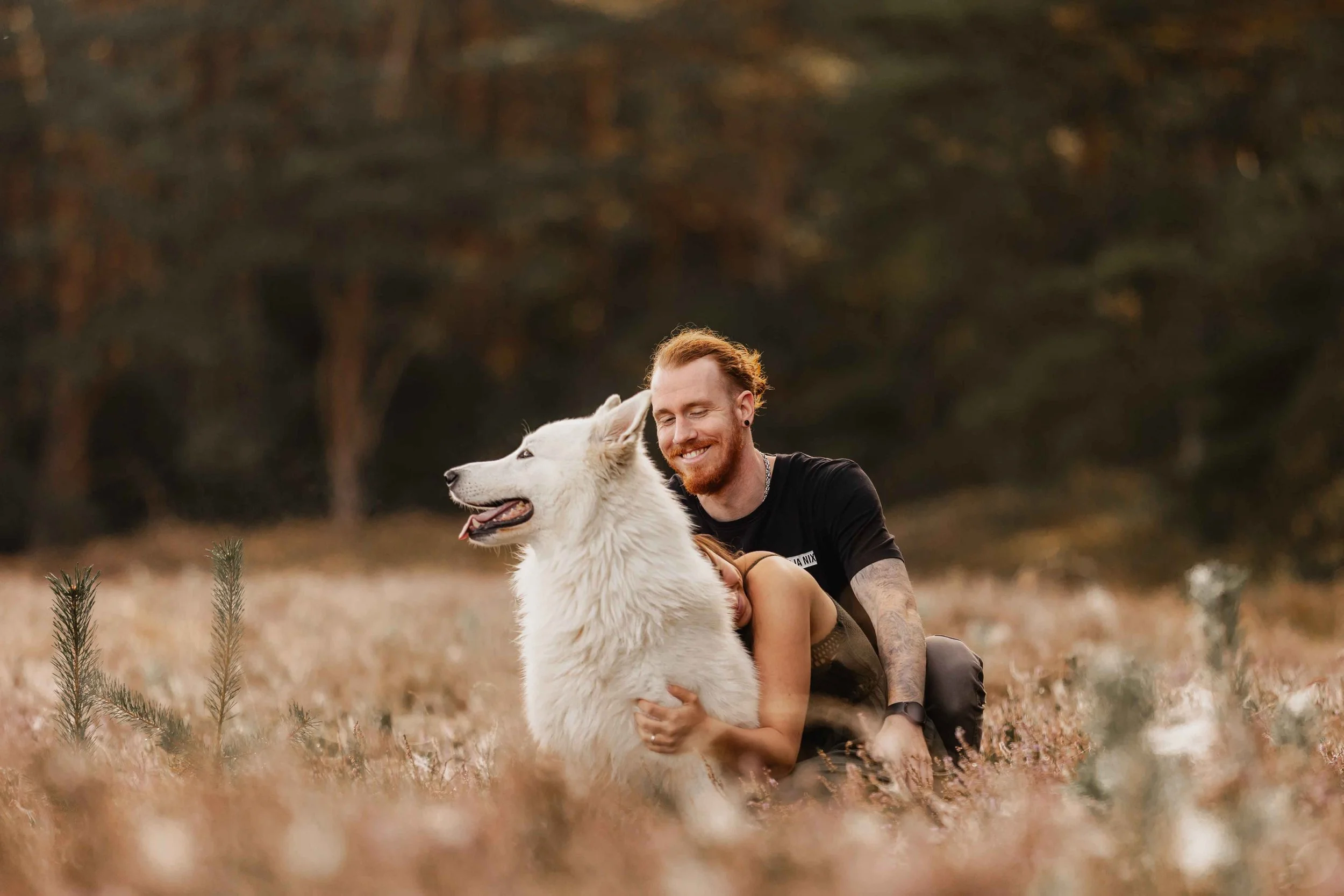Mann und Frau sitzen mit ihrem weißen Hund in der Heide, lachend und entspannt in der warmen Abendstimmung