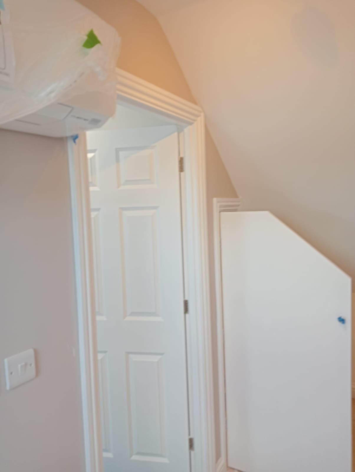 Part of an indoor room with a closed white door, beige walls, trim, and a sloped ceiling. A white plastic bag is hanging on the wall, and a small white cabinet is mounted on the wall.