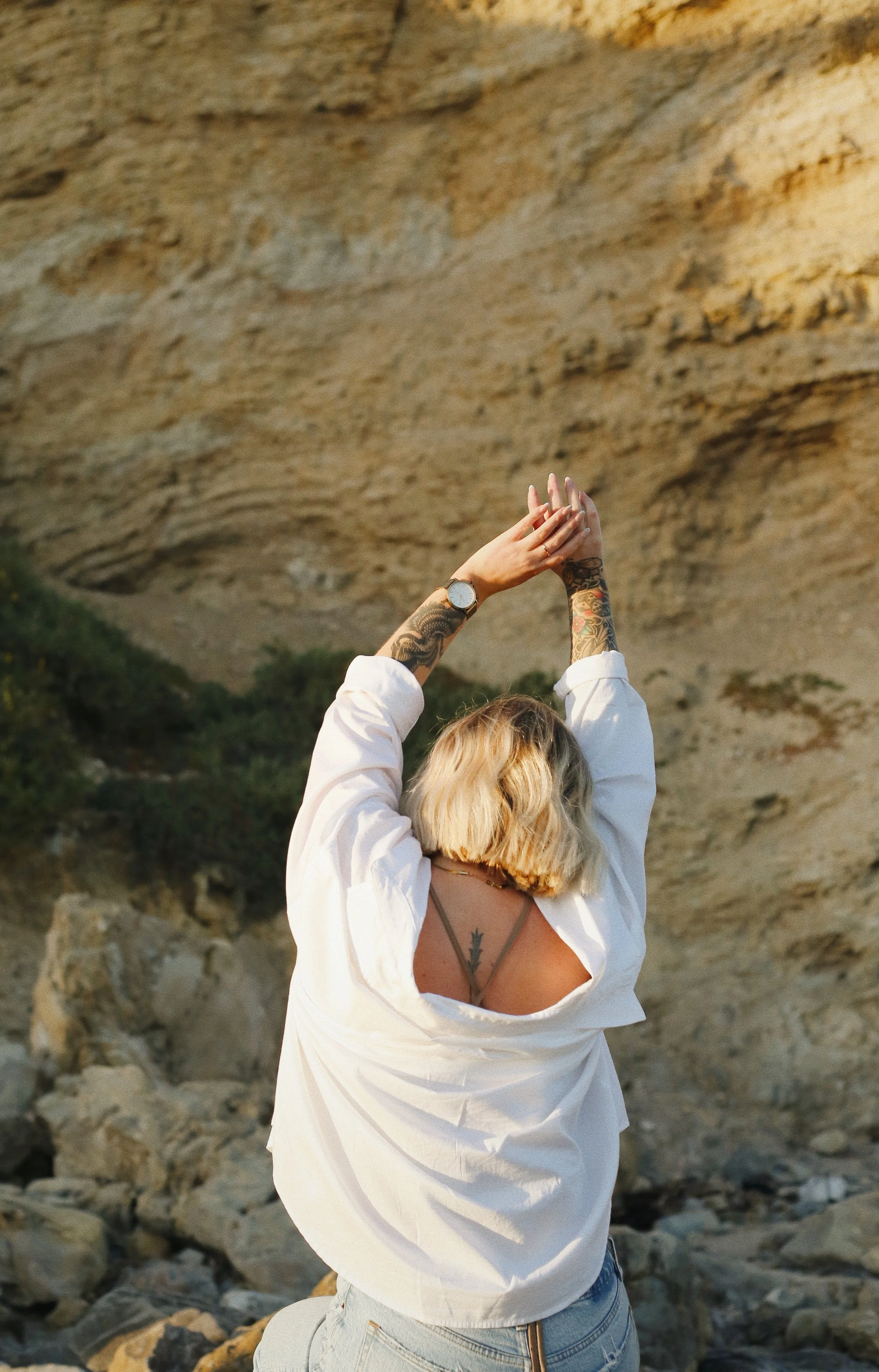Mujer con tatuajes en brazos y espalda, con blusa blanca y jeans, levantando los brazos en un entorno de rocas y acantilados durante el atardecer.