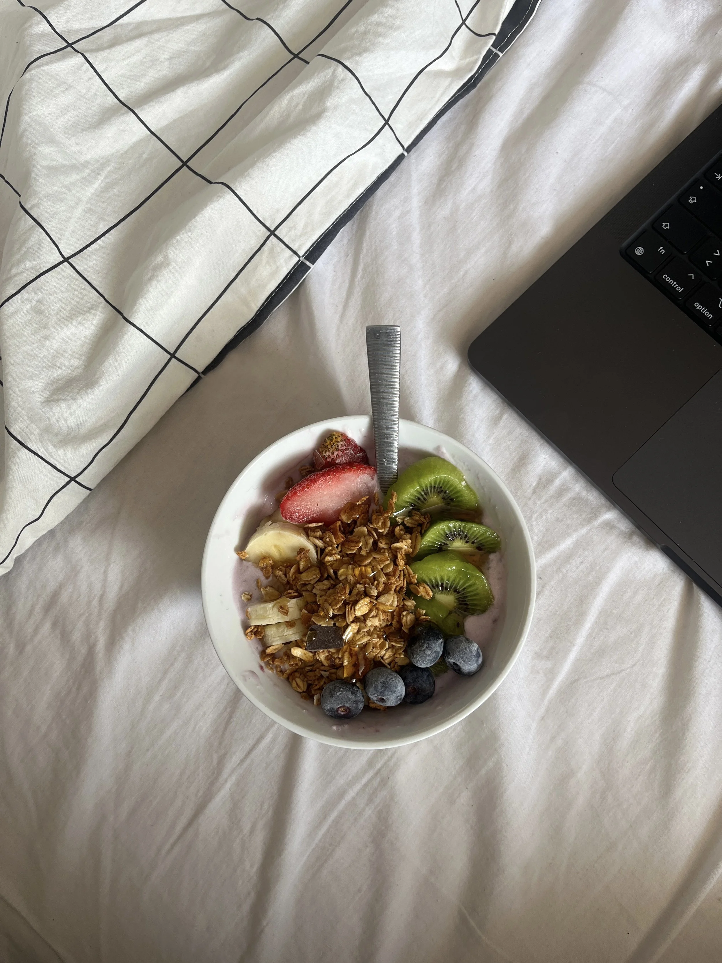 Cuenco de yogur con granola, fresas, plátano, kiwi y arándanos sobre una cama, con una cuchara metálica y un portátil negro a un lado.
