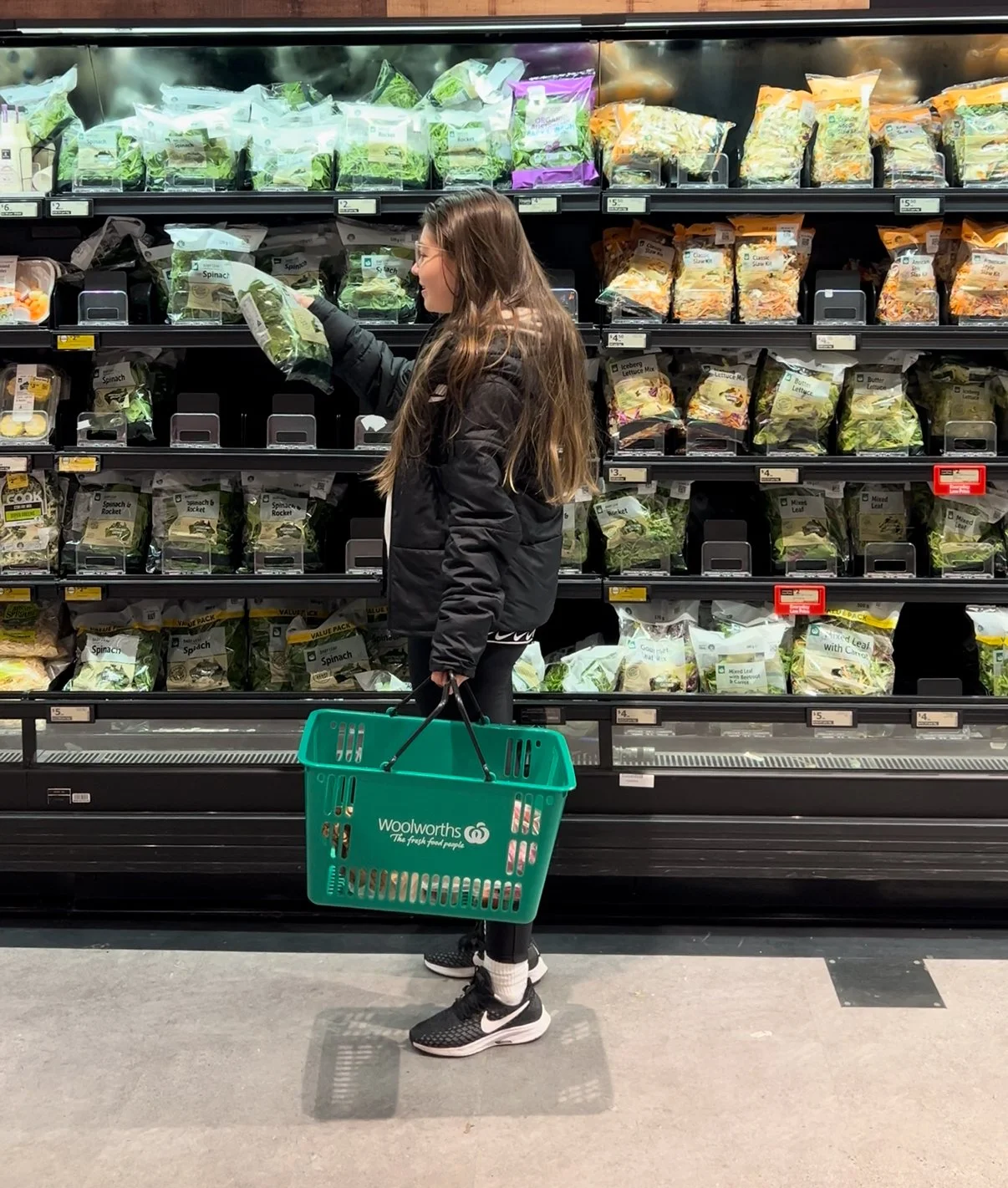 Luisa Cleves Nutricionista comprando ensaladas en el supermercado, sosteniendo una cesta de Woolworths, con estantes llenos de paquetes de verduras y ensaladas.