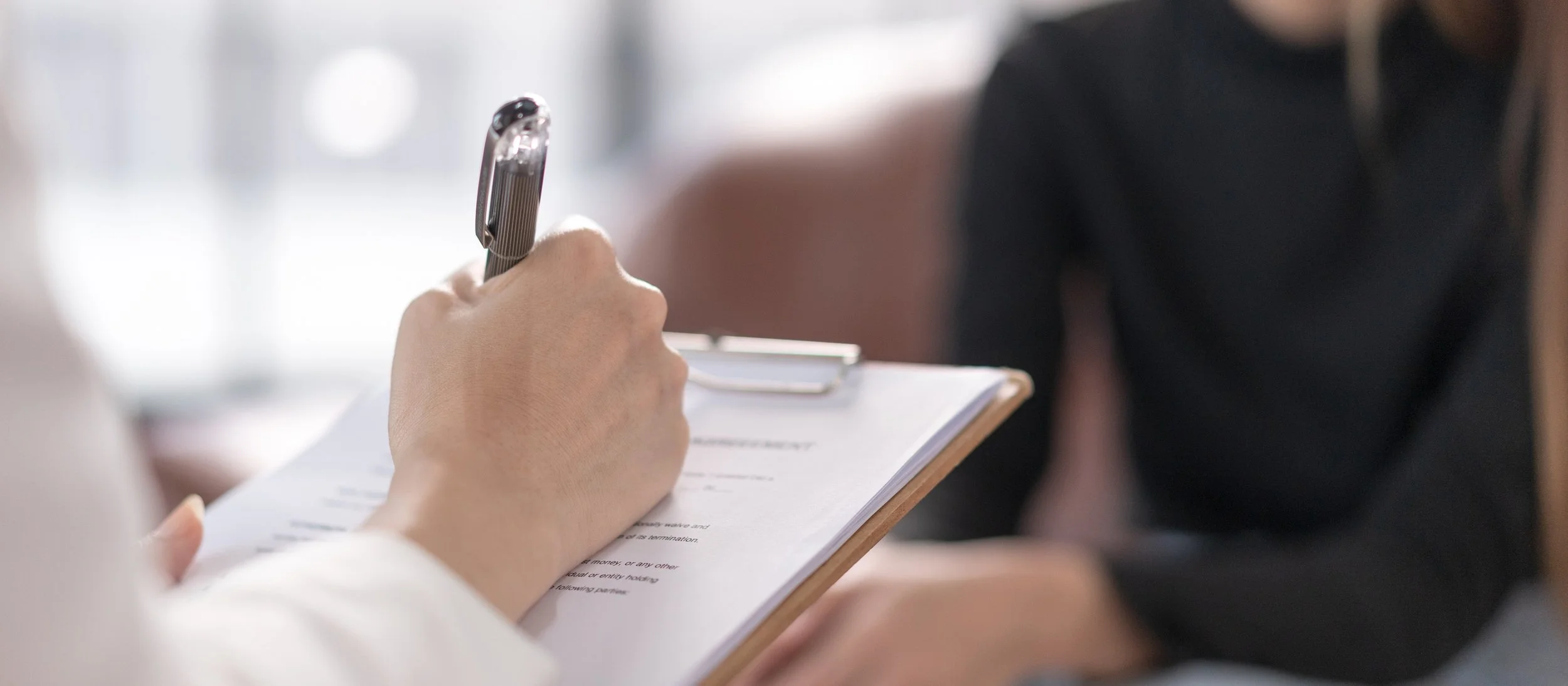 Person writing on a clipboard with a pen, with another person sitting nearby.