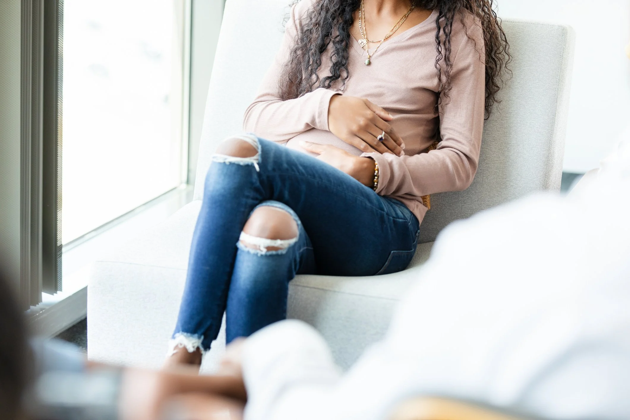 Woman sitting with crossed legs, wearing distressed jeans, a long-sleeve pink top, and jewelry near a window.