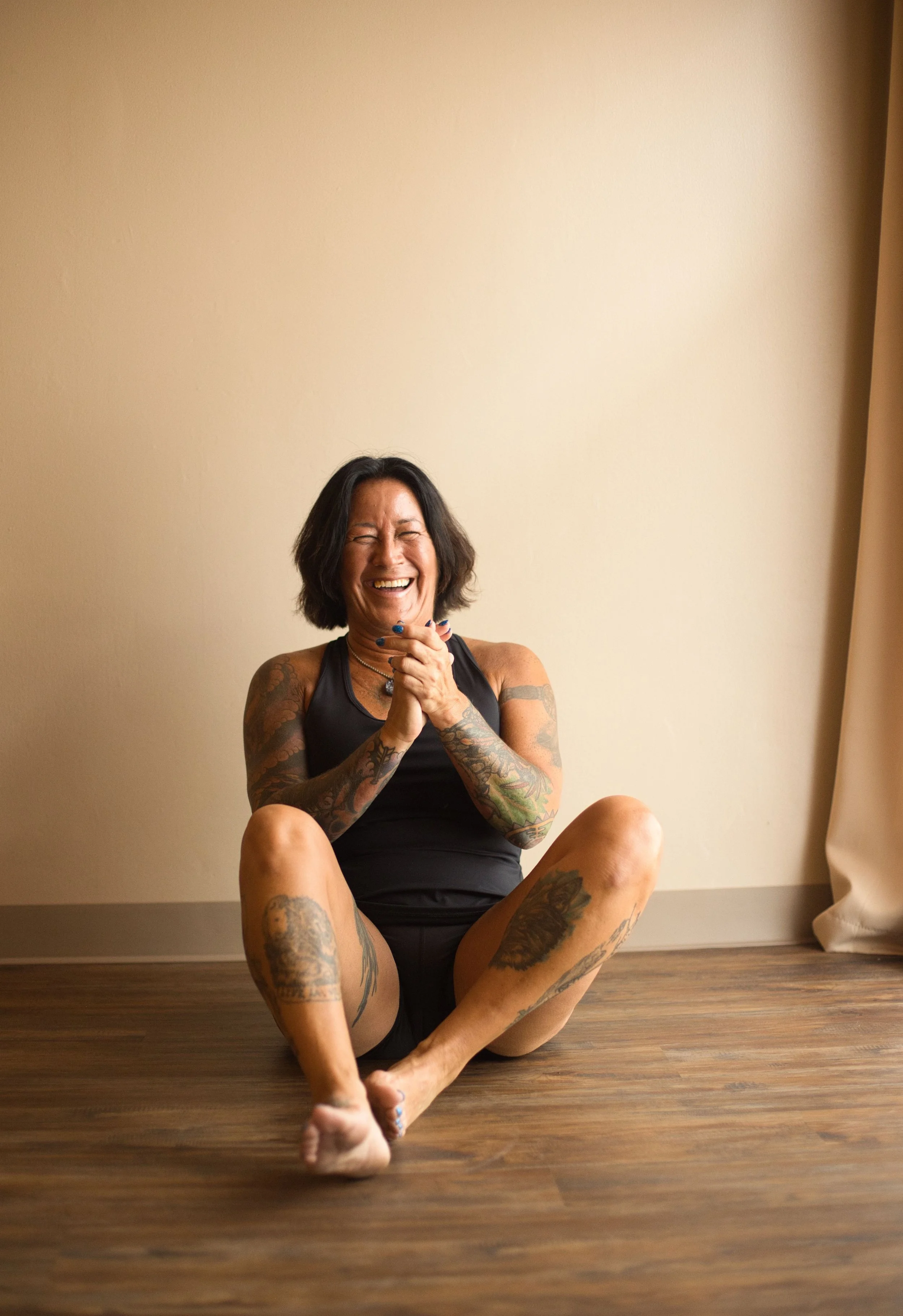A woman with short black hair, tattoos on her arms and legs, sitting on a wooden floor against a plain wall, smiling and laughing with her hands clasped together.