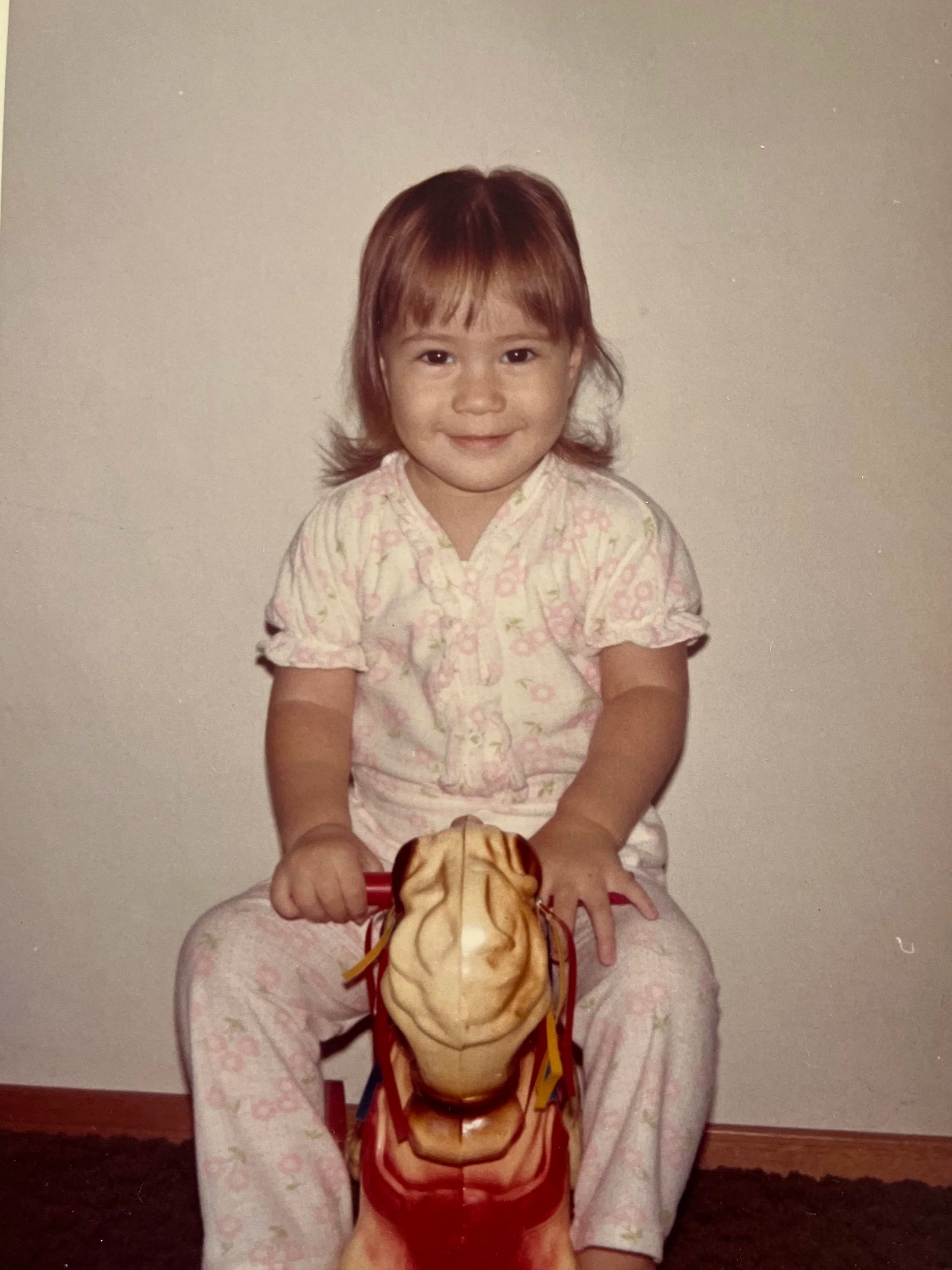 A young girl with brown hair sits on the floor wearing pink floral pajamas, smiling at the camera. She holds a skirt on her lap and a large toy head resembling a woman with long hair and a dress.