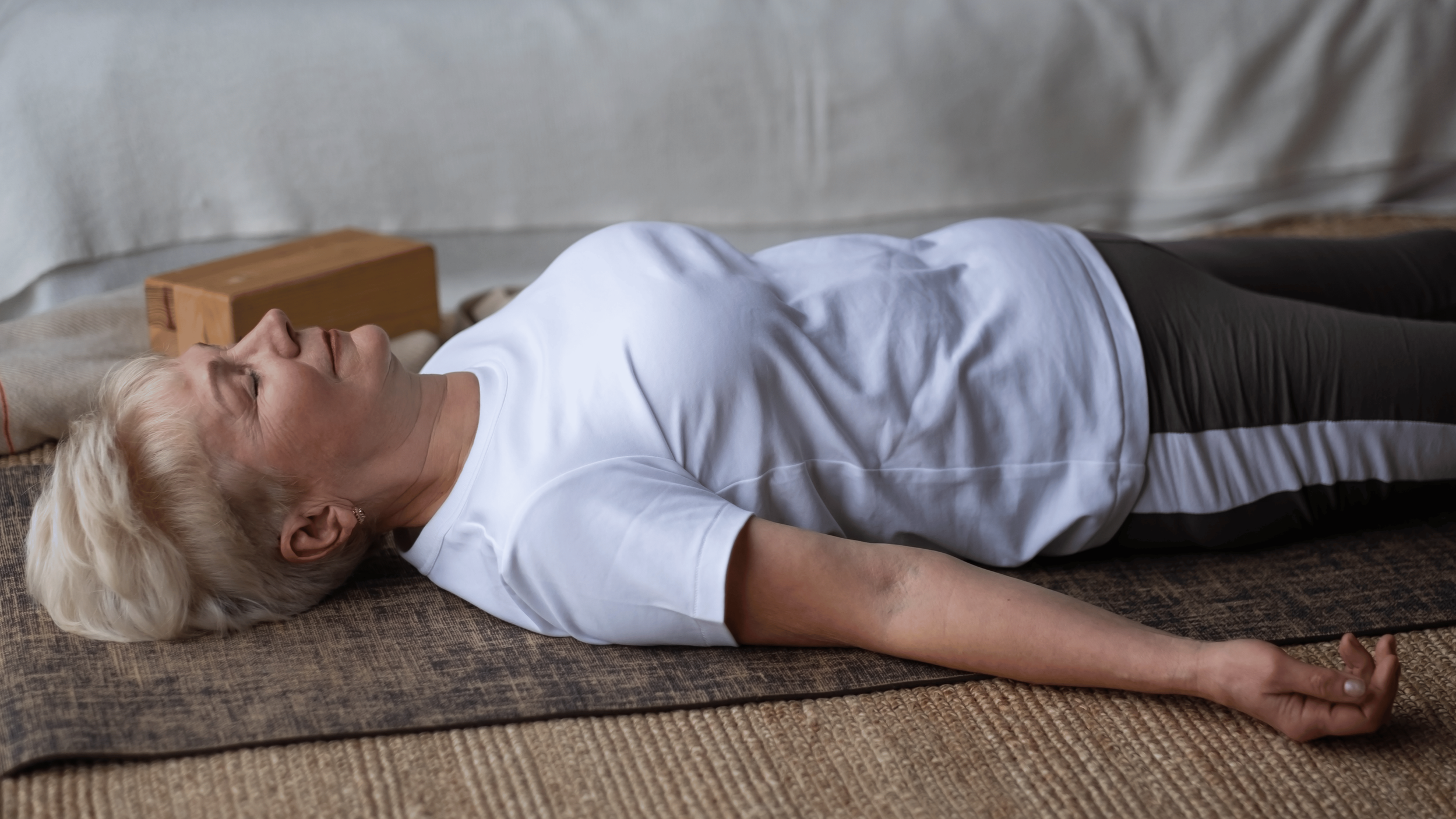 An older woman with blonde hair lying on her back on a mat, eyes closed, in a peaceful state, possibly practicing relaxation or meditation.