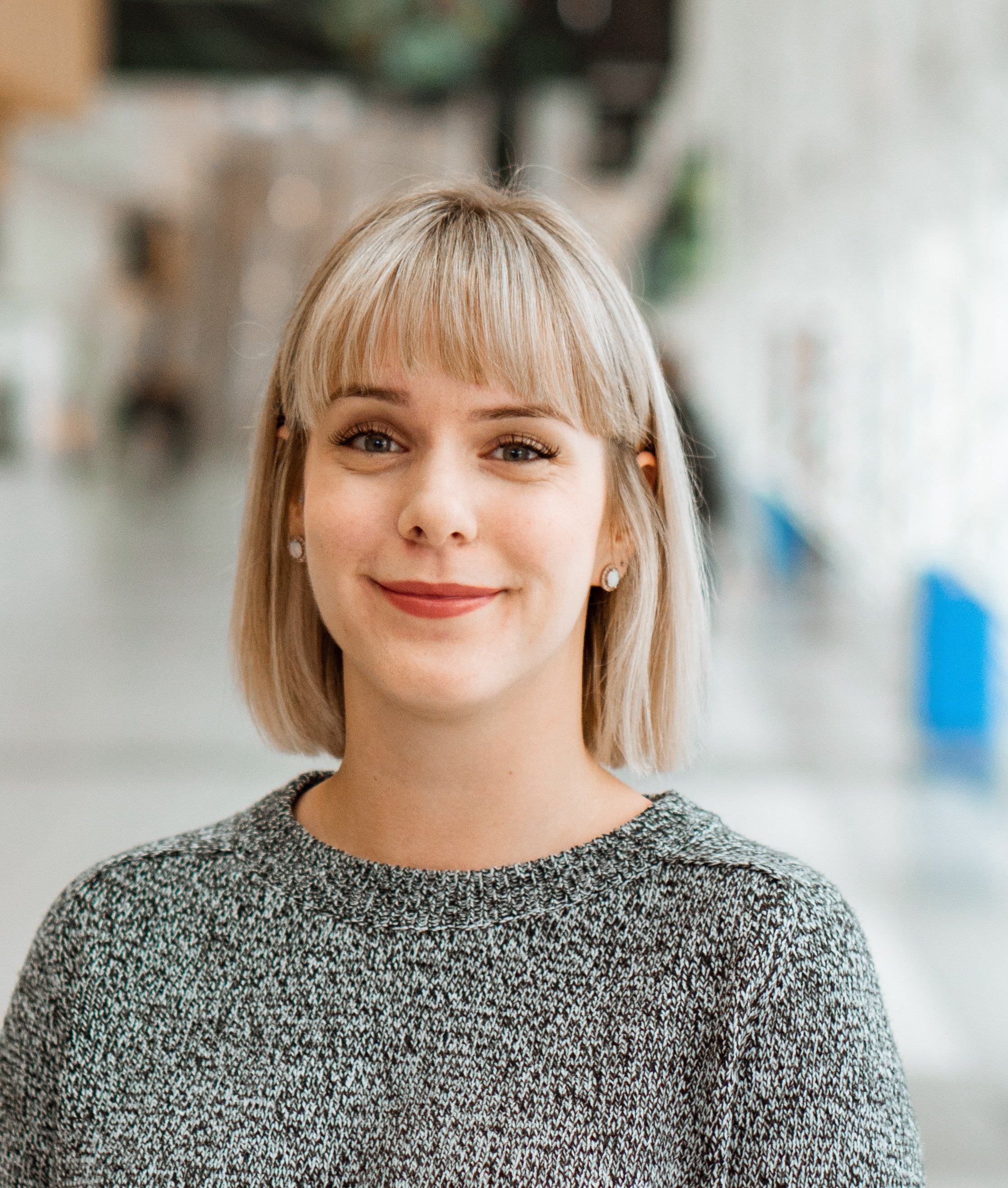 A young woman with blonde hair, wearing earrings and a grey sweater, smiling indoors.