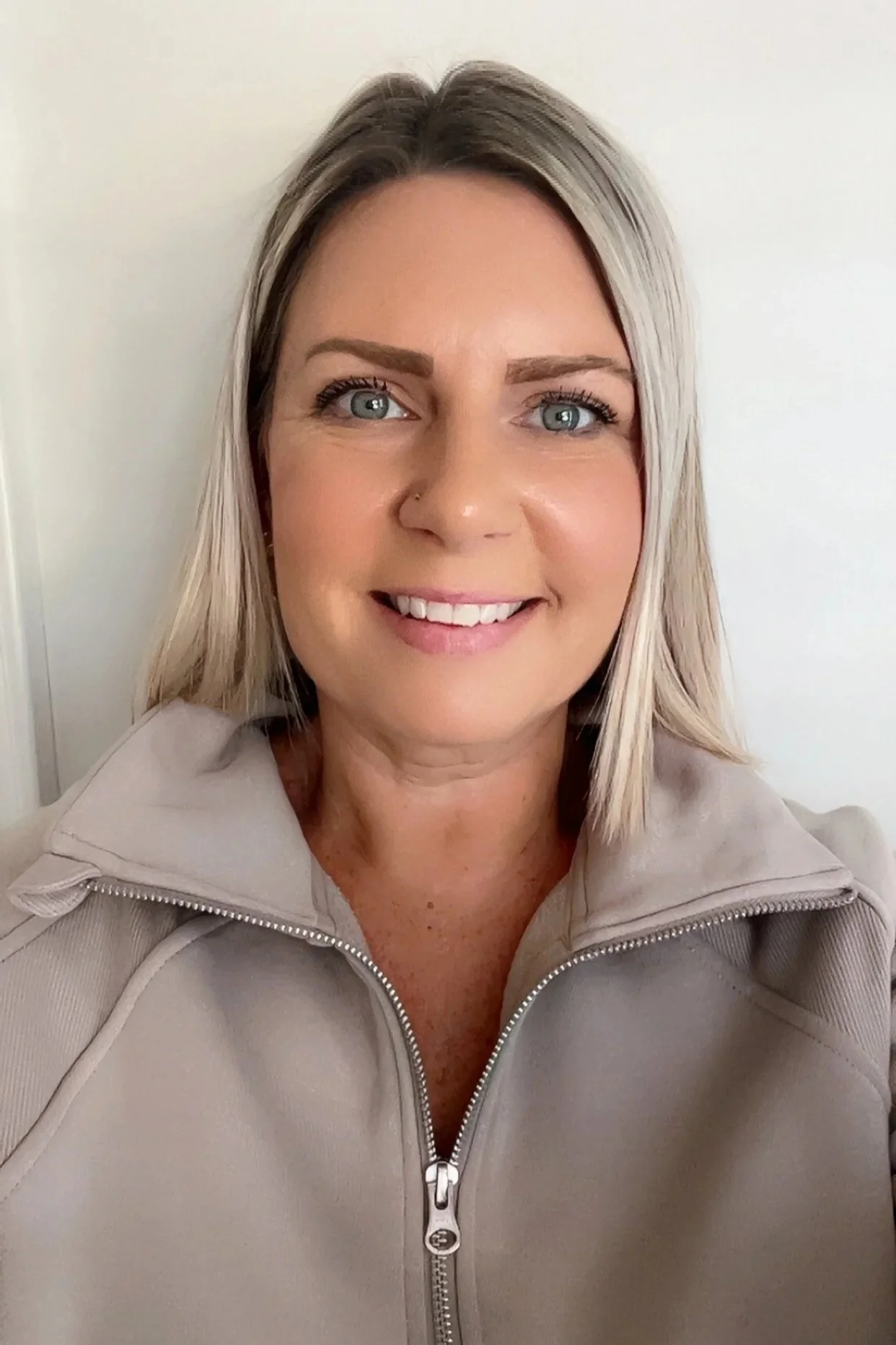 A woman with blonde hair smiling at the camera, wearing a light gray zip-up jacket with a neutral indoor background.