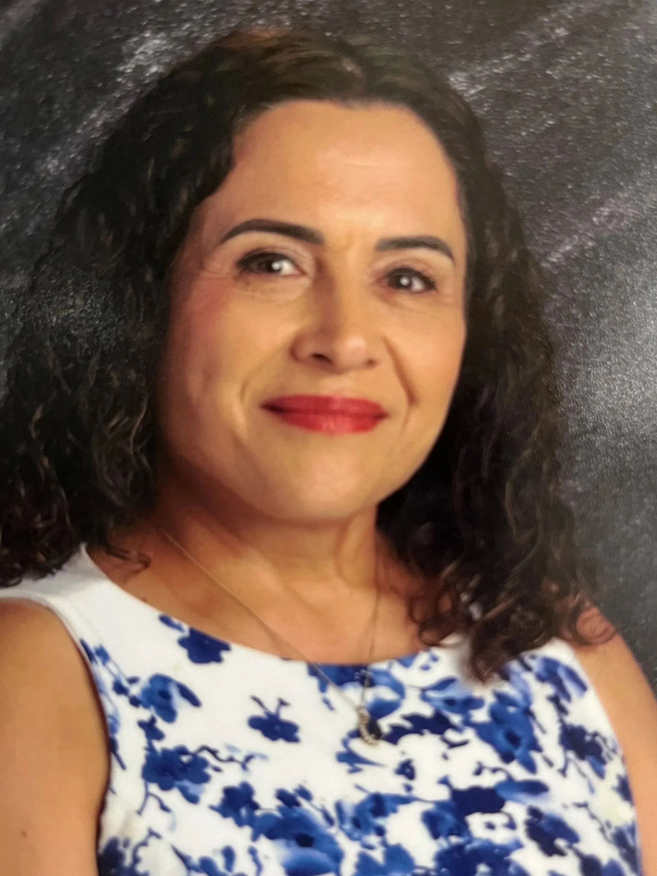 A woman with dark curly hair, wearing a sleeveless white dress with blue floral pattern, and red lipstick, smiling at the camera against a black textured background.