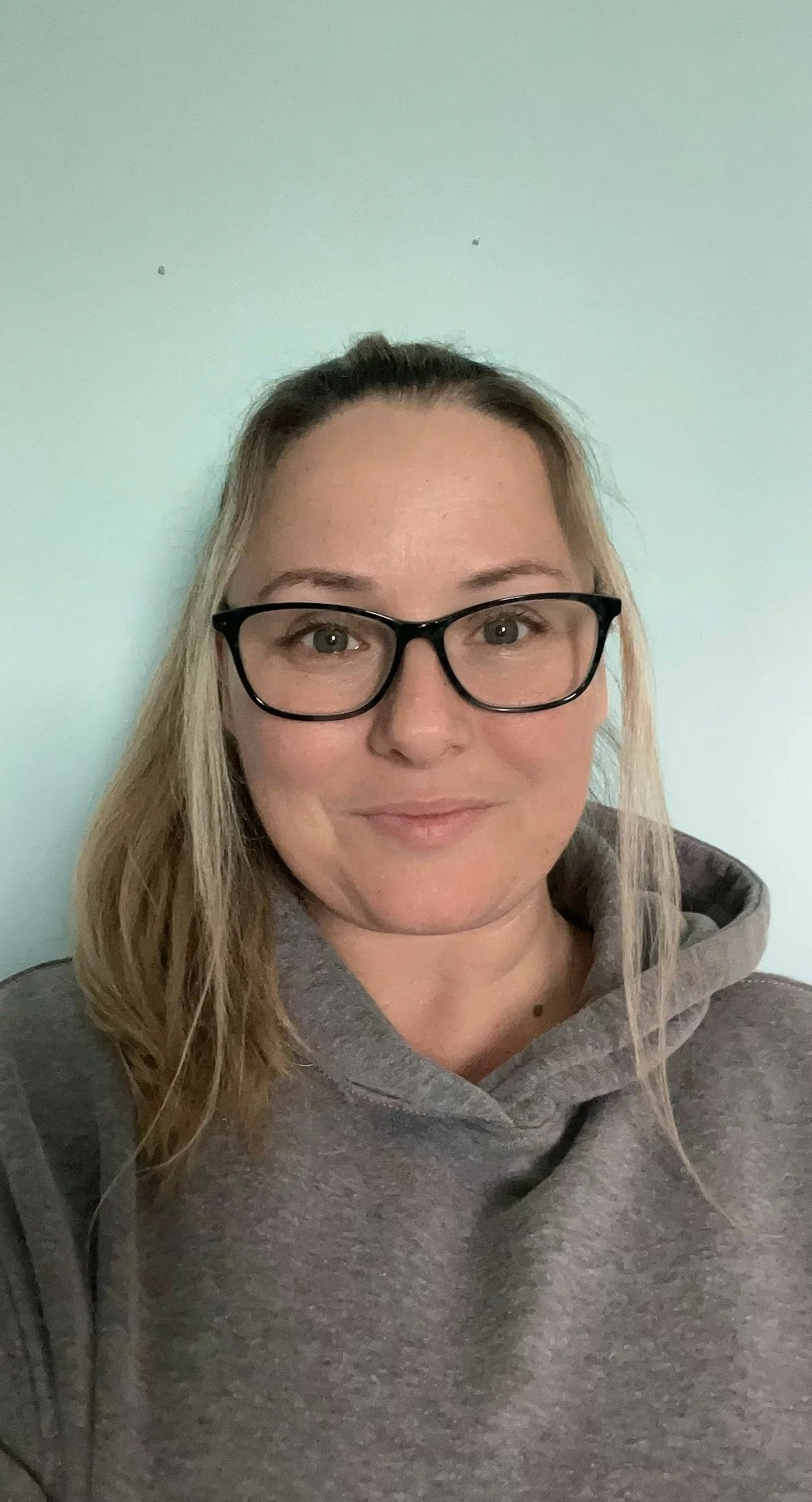 A woman with blonde hair, glasses, and a grey hoodie smiling at the camera against a pale green wall.