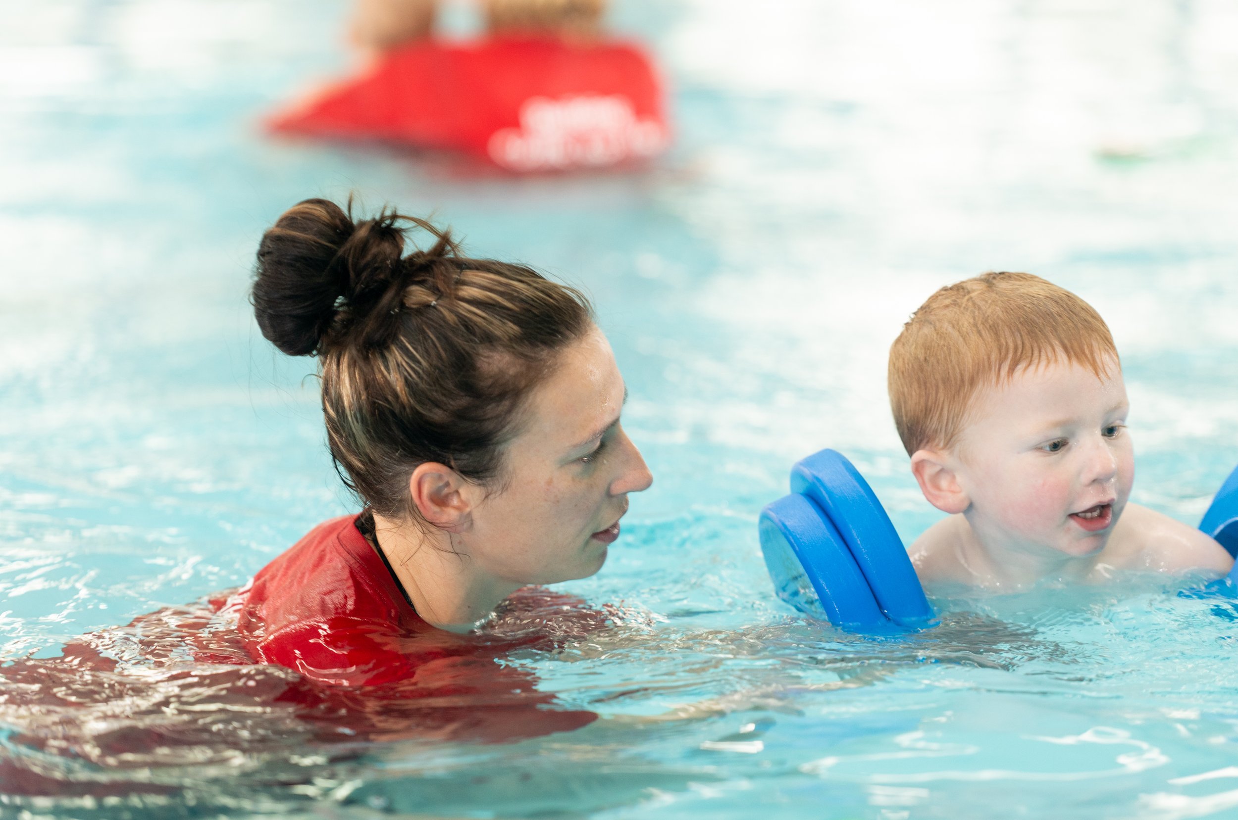 A woman with a bun hairstyle is helping a young red-haired boy learn to swim in a swimming pool. The boy is using blue foam arm floaties and is smiling.