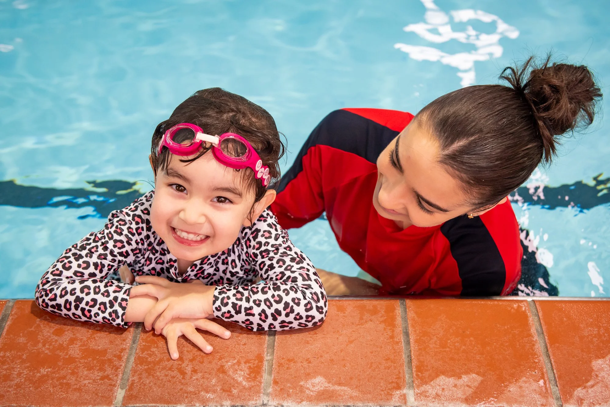 A young girl with pink swim goggles and a leopard print swimsuit smiling at the camera while leaning on the edge of a swimming pool, with a woman beside her holding her in the water.