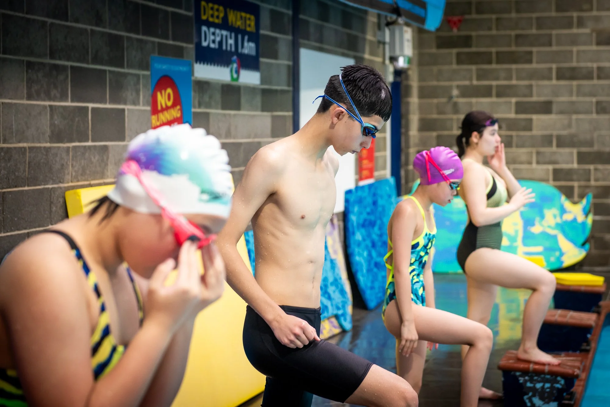 Swimmers preparing for practice in an indoor pool area, wearing swim caps and goggles, with a wall and signs in the background.