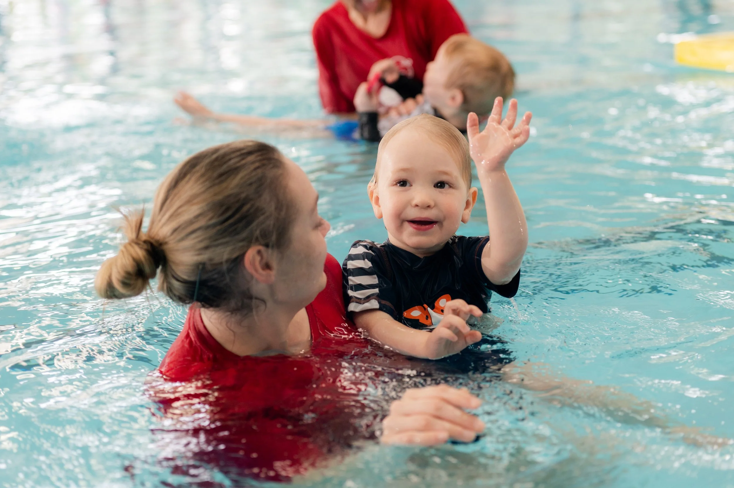 A smiling toddler waving in a swimming pool while being held by an adult woman, with other people in the background in the water.